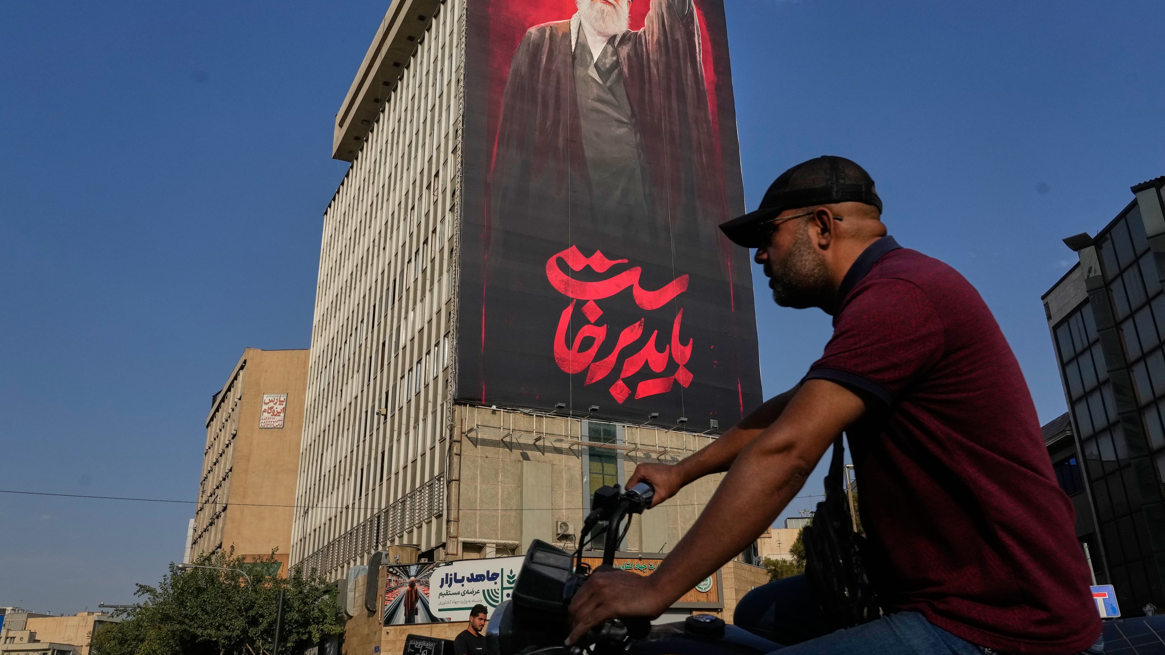 A man rides on his motorbike past a billboard with a message that reads in Farsi: " We must rise up" and an image of the late Iranian Supreme Leader Ayatollah Ali Khamenei, who was killed during the U.S. and Israel strikes on Feb. 28, in Tehran, Iran, Saturday, Aug. 15, 2026. (AP Photo/Vahid Salemi)