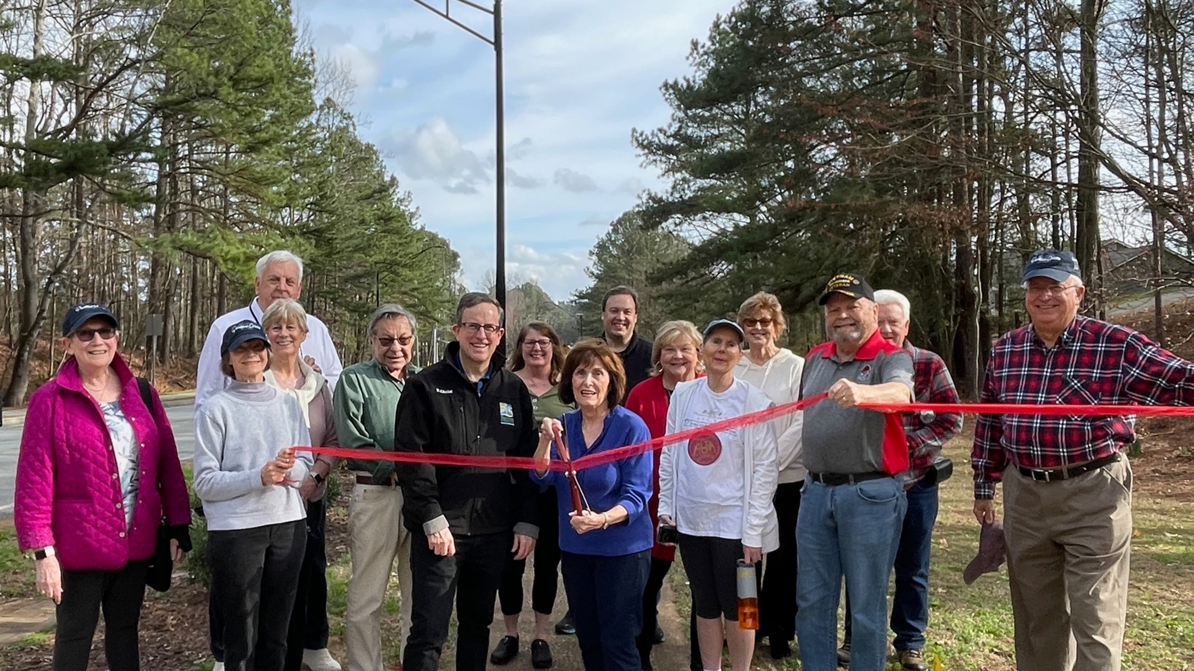 Susan Albright (with scissors) and Peachtree Corners Councilmember Eric Christ (in black jacket), along with Dunwoody Manor neighbors, ready to cut the ribbon with one of the new streetlights along Jay Bird Alley in the background. (Courtesy City of Peachtree Corners)