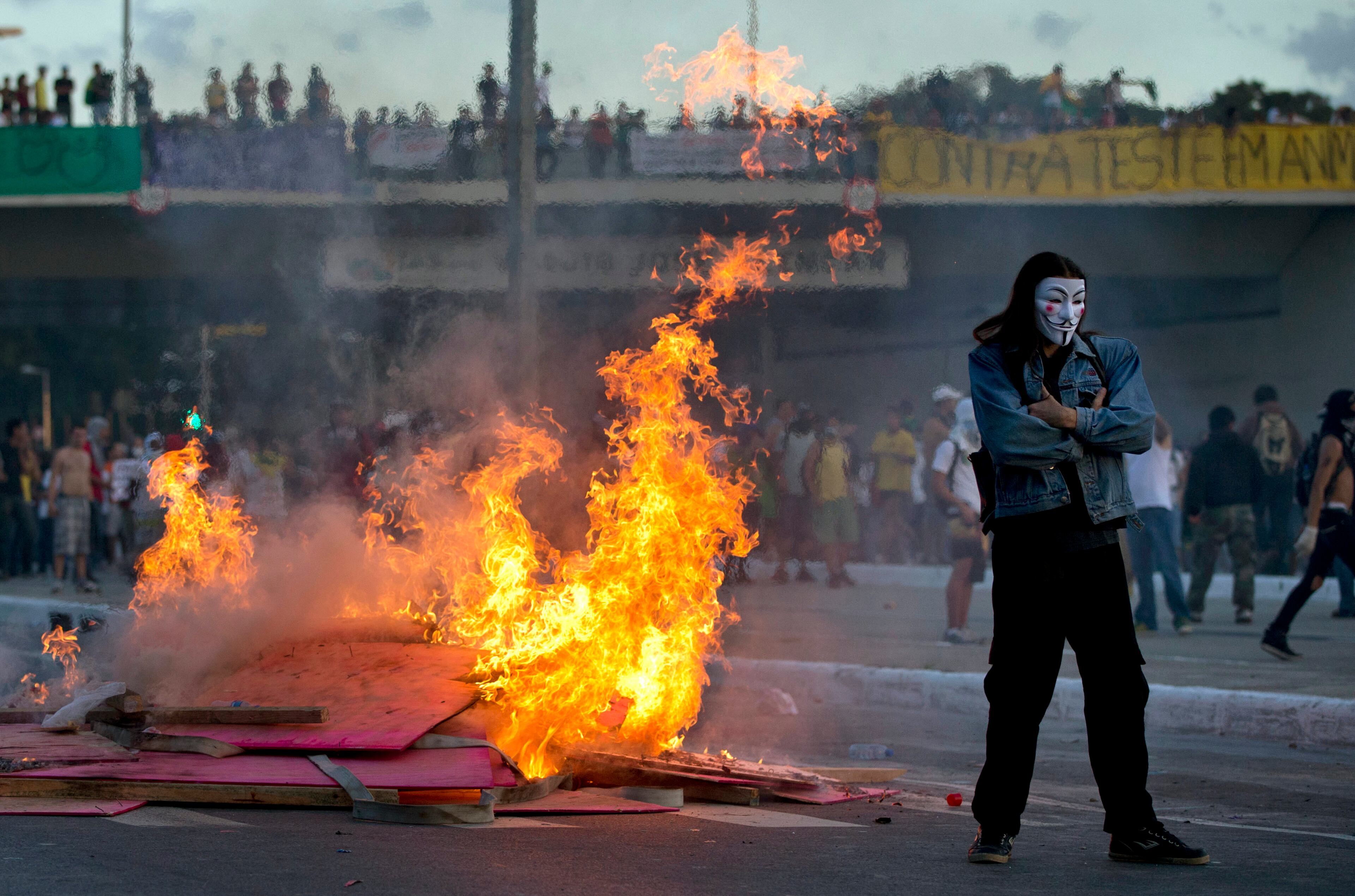 A protester with a mask stands by a burning barricade during a demonstration in Belo Horizonte, Brazil, Wednesday, June 26, 2013. Brazilian anti-government protesters in part angered by the billions spent in World Cup preparations and police clashed Wednesday near the stadium hosting a Confederations Cup football match, with tens of thousands of demonstrators trying to march on the site confronting police firing tear gas and rubber bullets.(AP Photo/Victor R. Caivano)