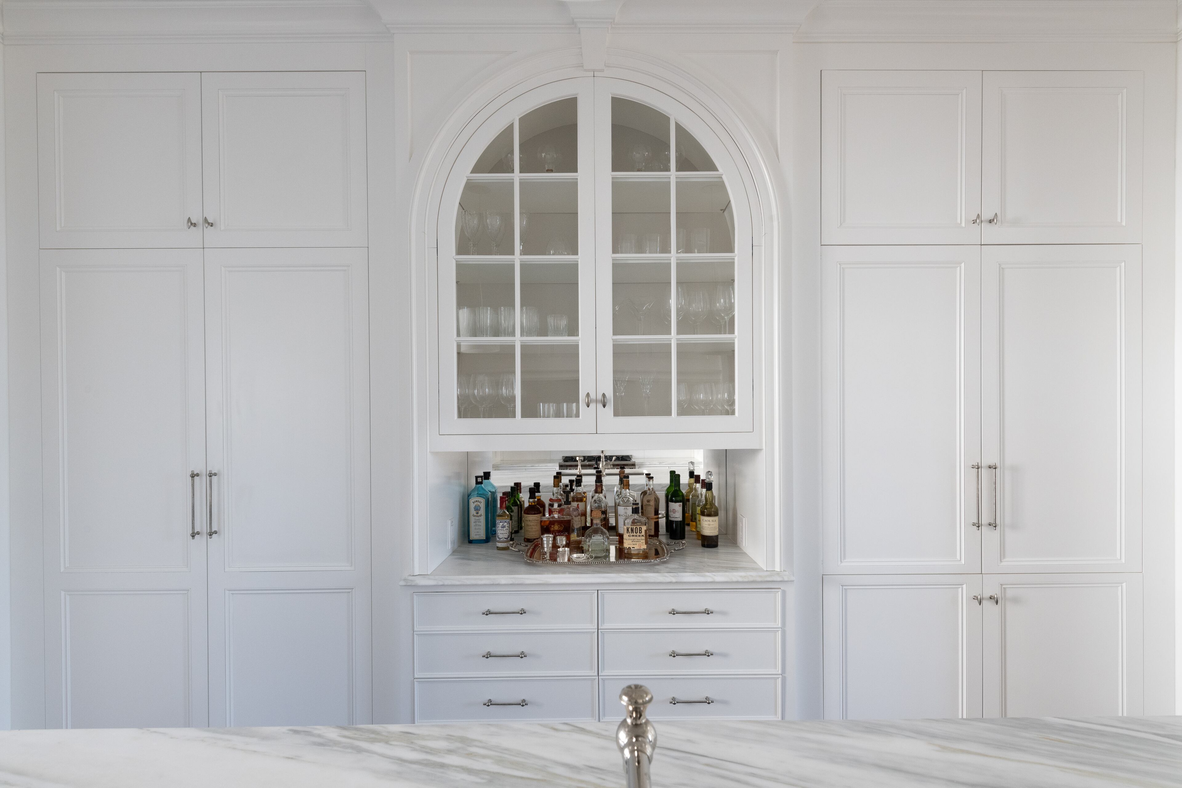 Mimicking the Roman arch motif found throughout the home, the bar's glass-front display cabinet doors create variety amongst the linearity of the kitchen design. Text by Ansley Warren