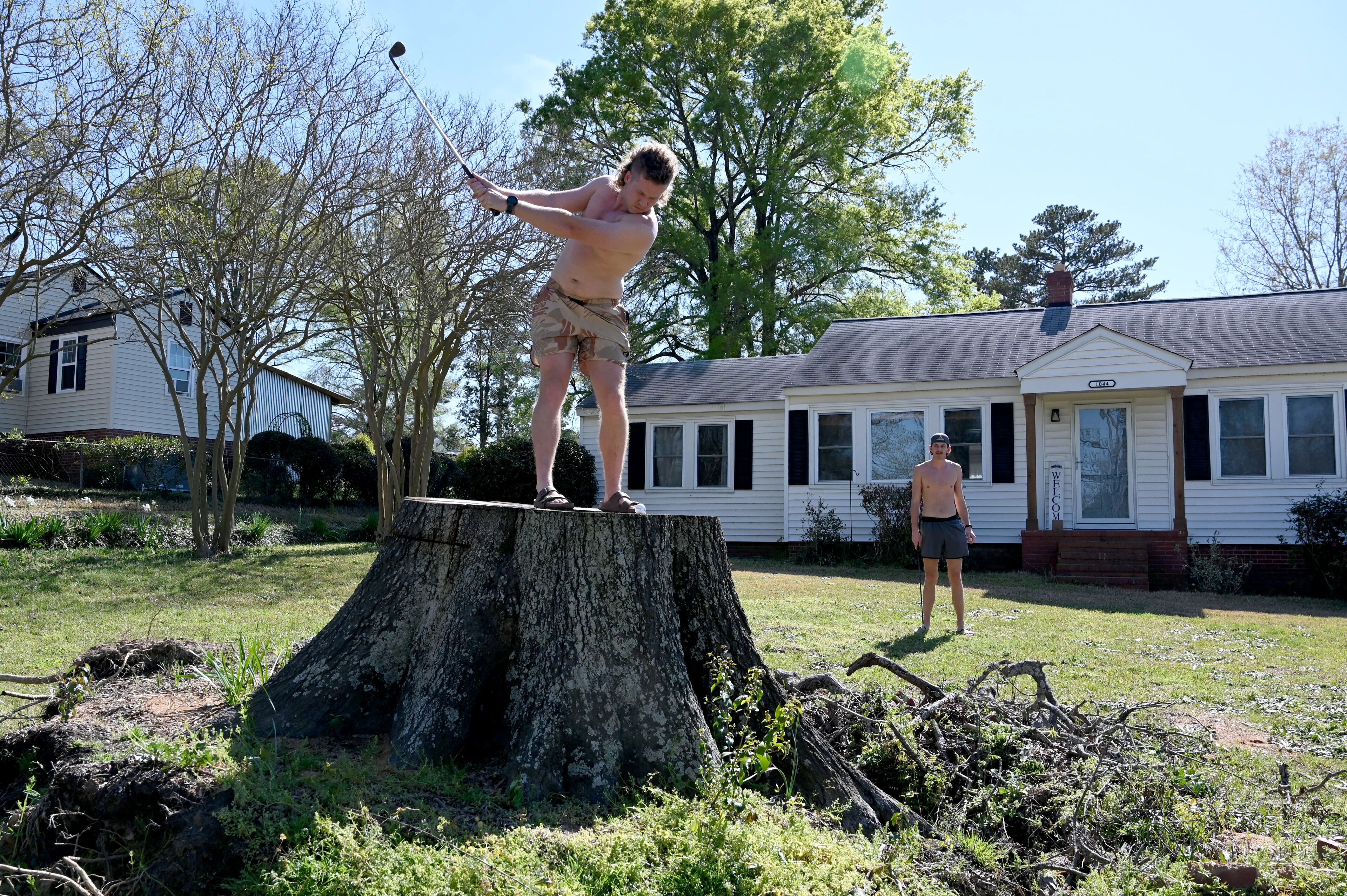 Obie Brannon hits a shot from the downed tree stump from Hurricane Helene as Walt Steward, both students at Dental College of Georgia, looks on at their front yard located within a mile away from Augusta National Golf Club, Tuesday, March 25, 2025, in Augusta. Six months after Hurricane Helene, Augusta-area community is still working to recover from the extensive damage. (Hyosub Shin / AJC)