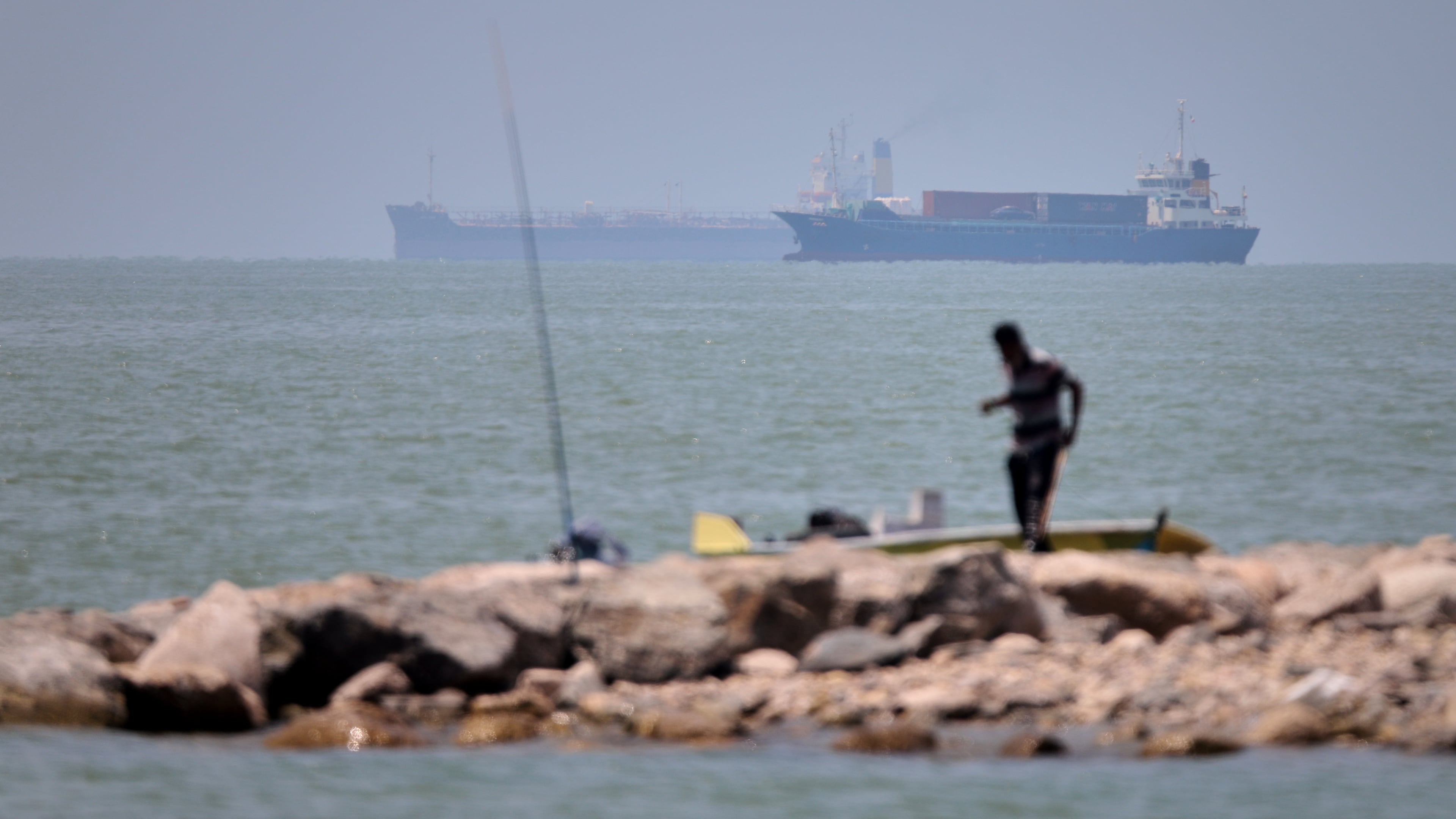 A man stands beside a fishing pole along the shore as cargo ships and commercial vessels are seen in the Strait of Hormuz off Bandar Abbas, Iran, Wednesday, June 17, 2026. (Amirhosein Khorgooi/ISNA via AP)