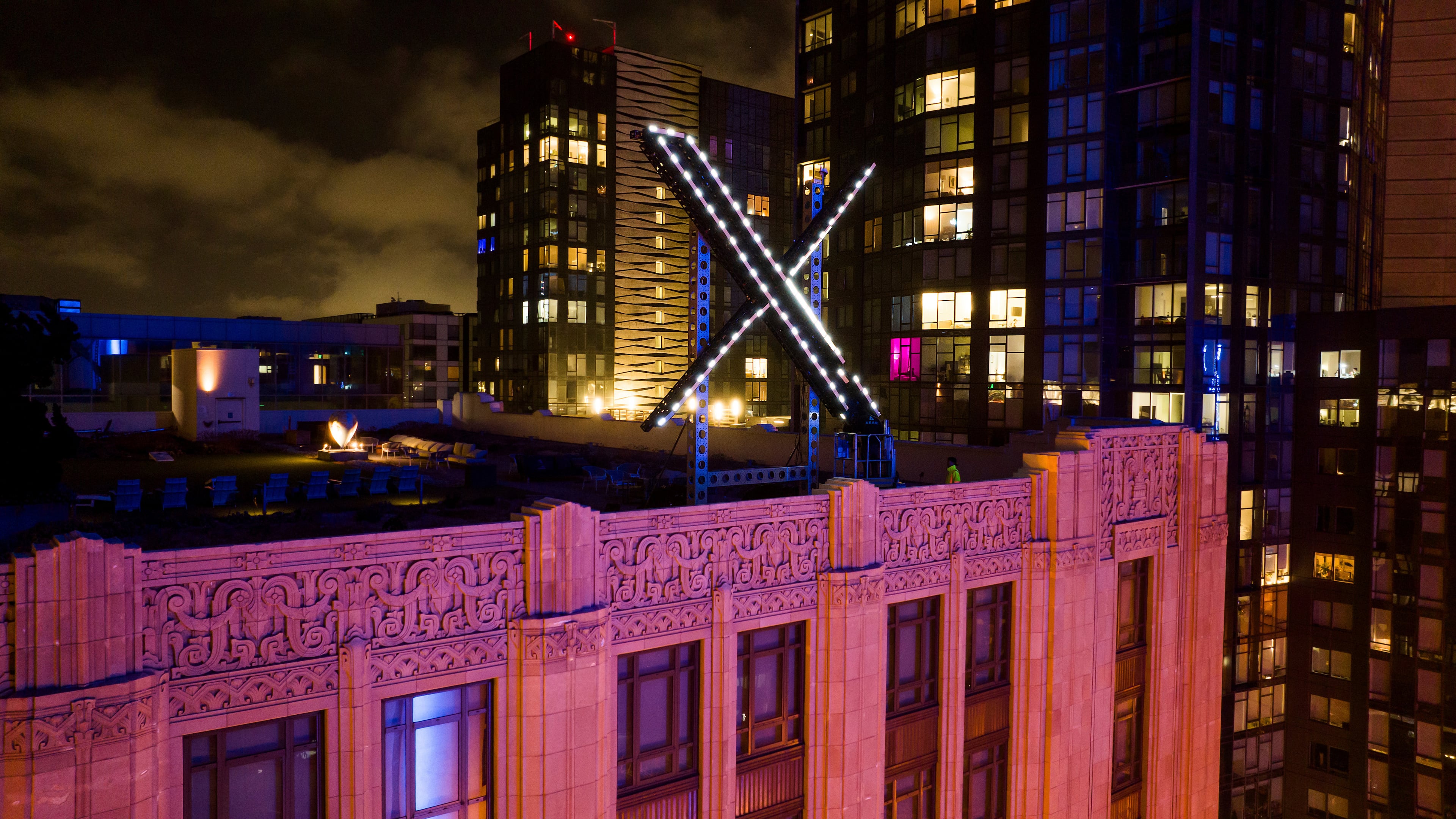 Workers install lighting on an "X" sign atop the company headquarters formerly known as Twitter in downtown San Francisco. X is owned by Elon Musk, who also owns the AI tool Grok, which has been scrutinized for its unrestricted creation of AI-generated nude images, including of adolescents and children. (AP 2023)