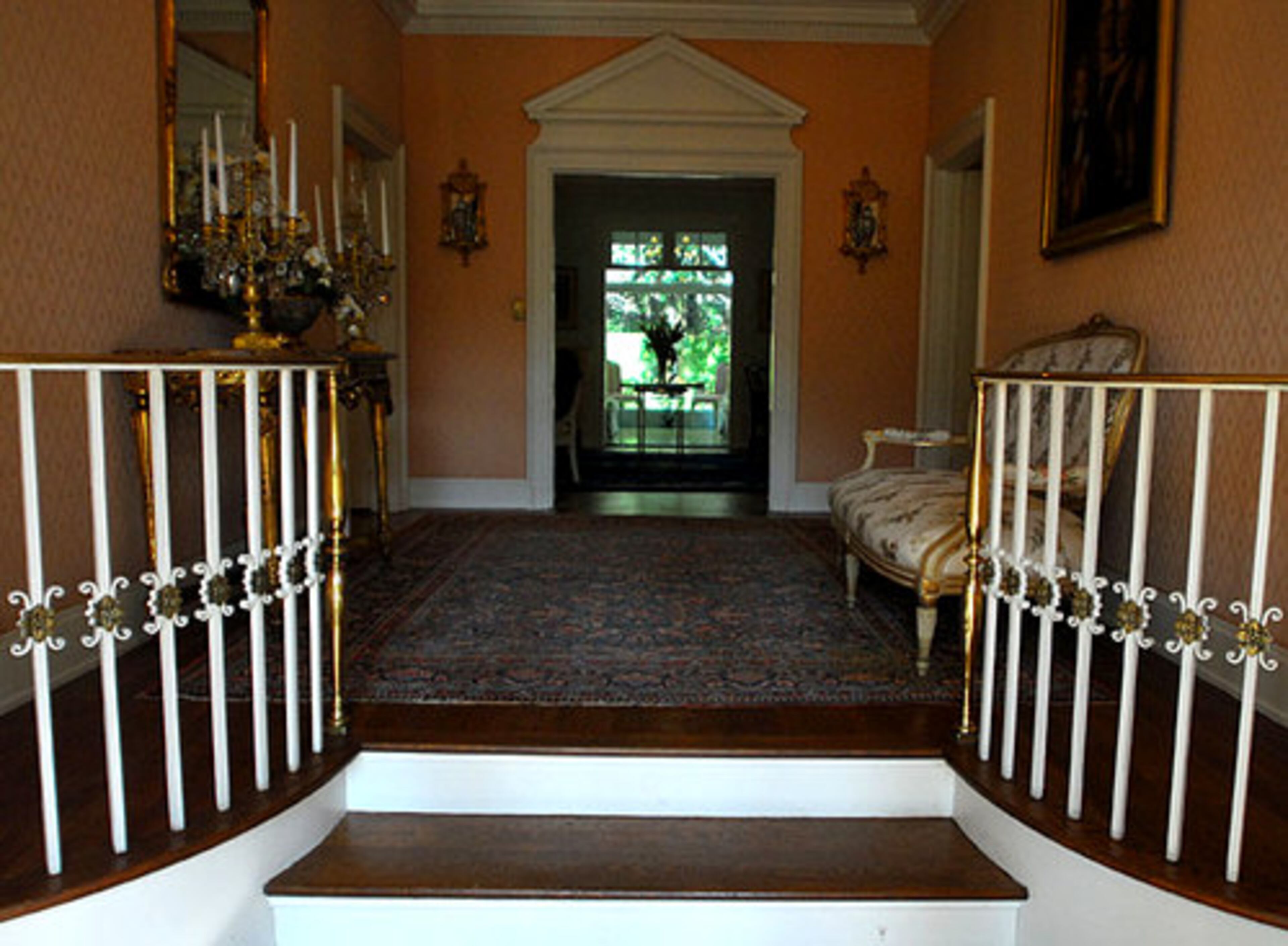 A brass rail lines the steps up into the home's foyer from the front door. The door separating the foyer from the living room echoes the classical motif of the front portico. Family portraits include Oakland Plantation owner Alfred Wellborn and his daughter, Mary Jane Wellborn Cochran.