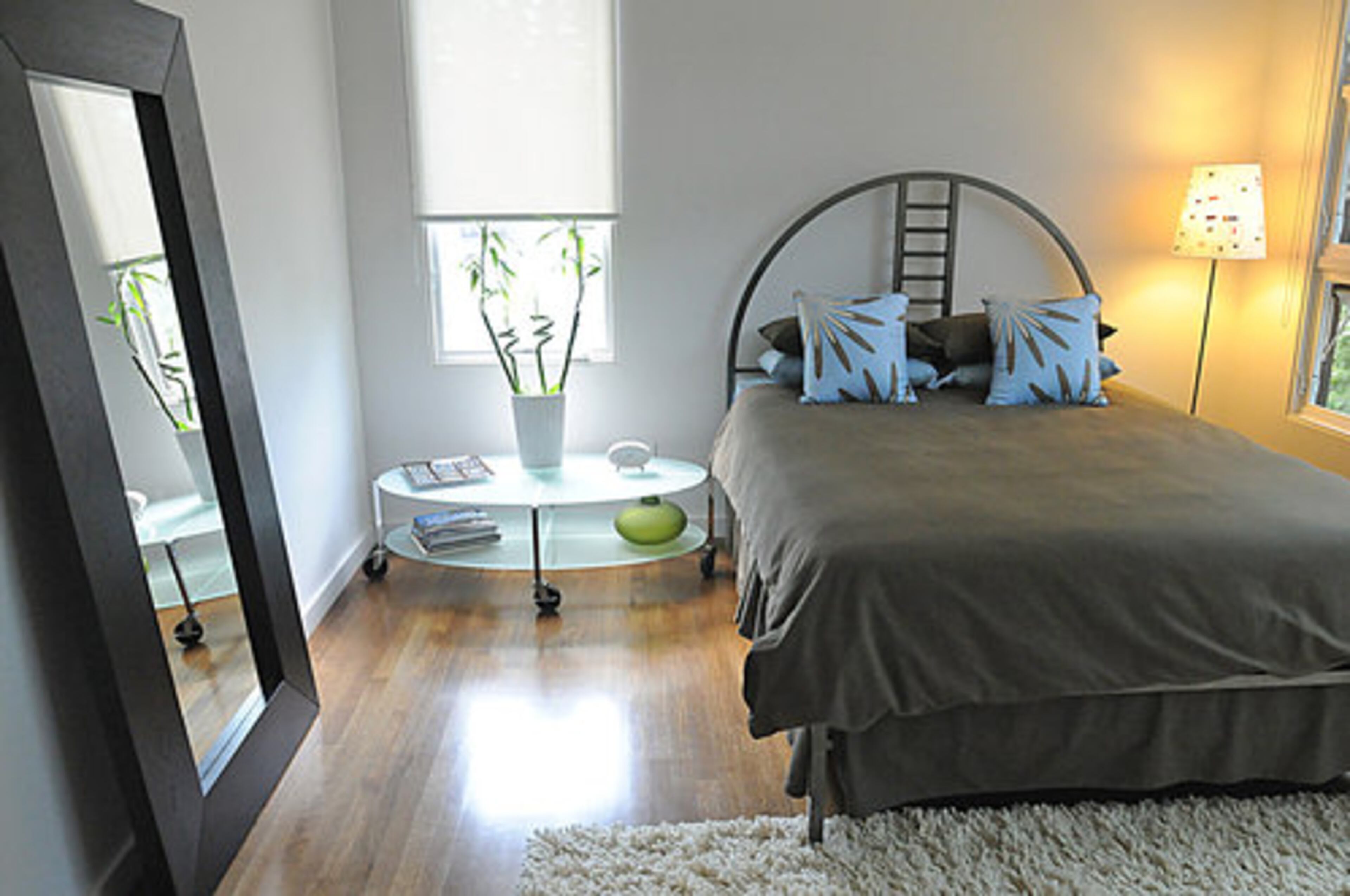 This bedroom has a cleverly placed mirror to maximize the space. The headboard of the bed features a unique ladder pattern in the center. Details like the decorative plate on the see-through nightstand table which complements the color scheme of the bedding are thoughtful touches.