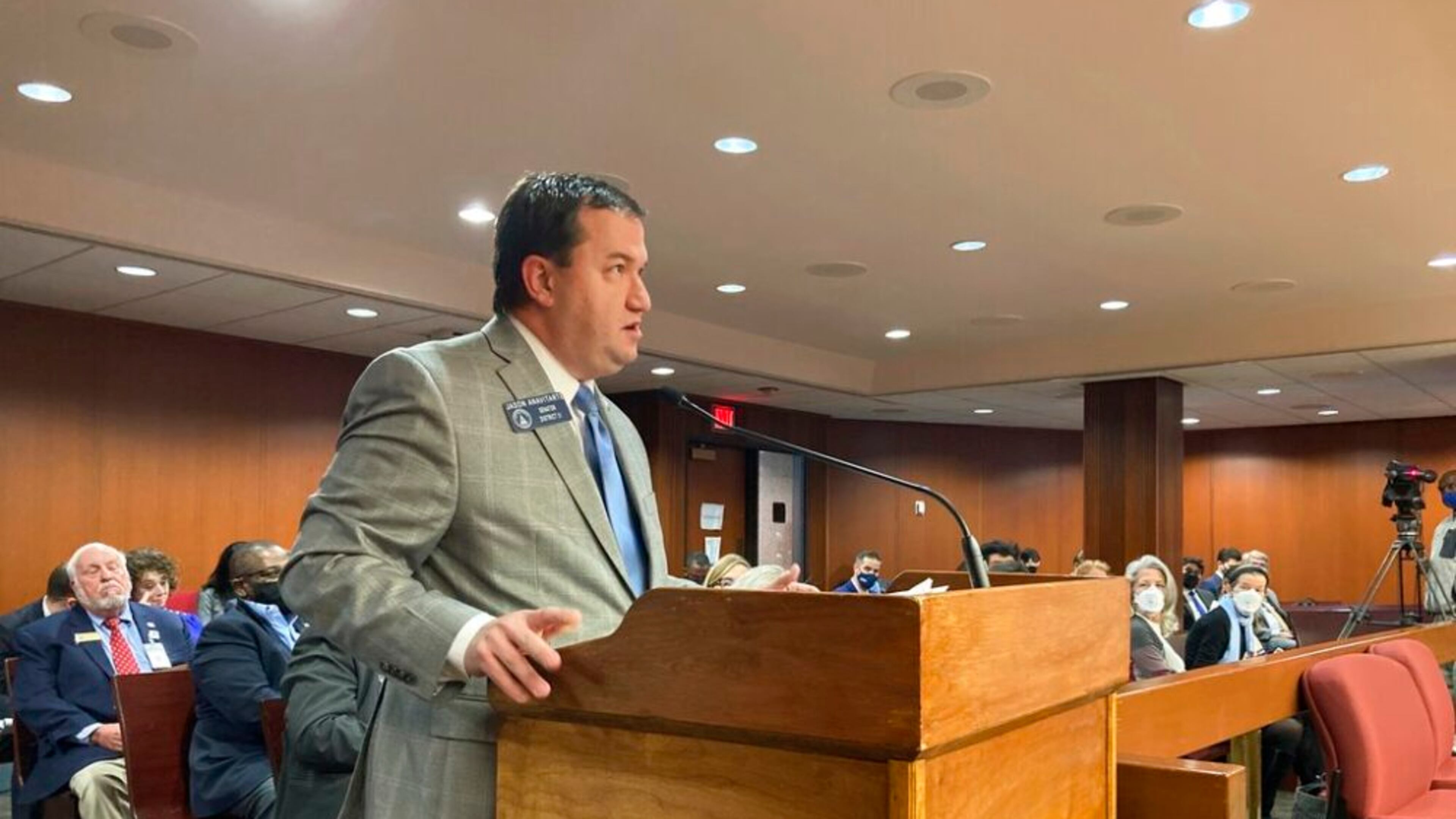white man with dark black hair, speaking into brown podium with grey blazer, blue pants and tie.