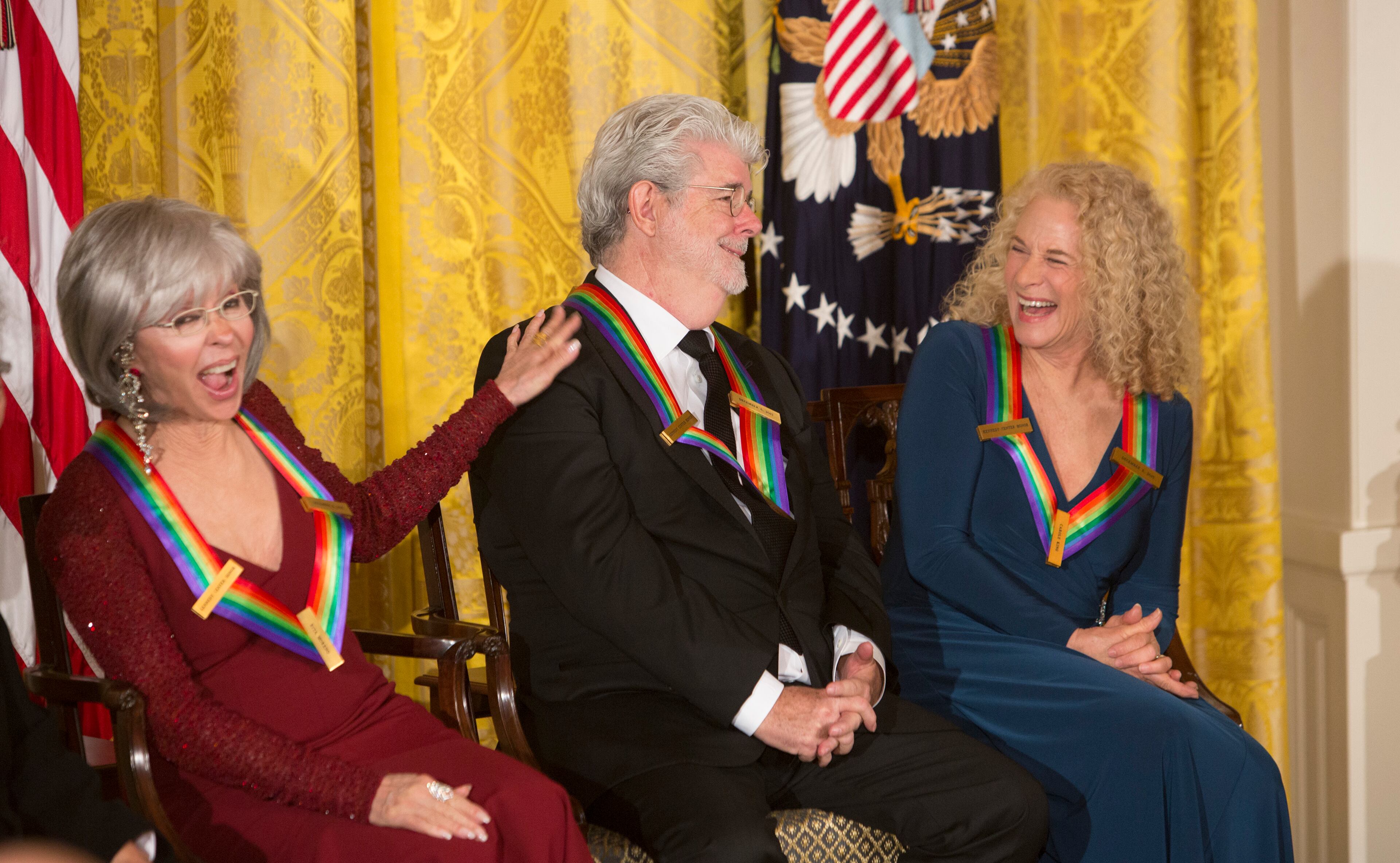 Actress and singer Rita Moreno (L) and singer-songwriter Carole King (R) laugh during the introduction of filmmaker George Lucas (C) as 2015 Kennedy Center Honors recipients, December 6, 2015 during a reception at The White House in Washington DC. AFP Photo / Chris Kleponis / AFP / CHRIS KLEPONIS (Photo credit should read CHRIS KLEPONIS/AFP/Getty Images)