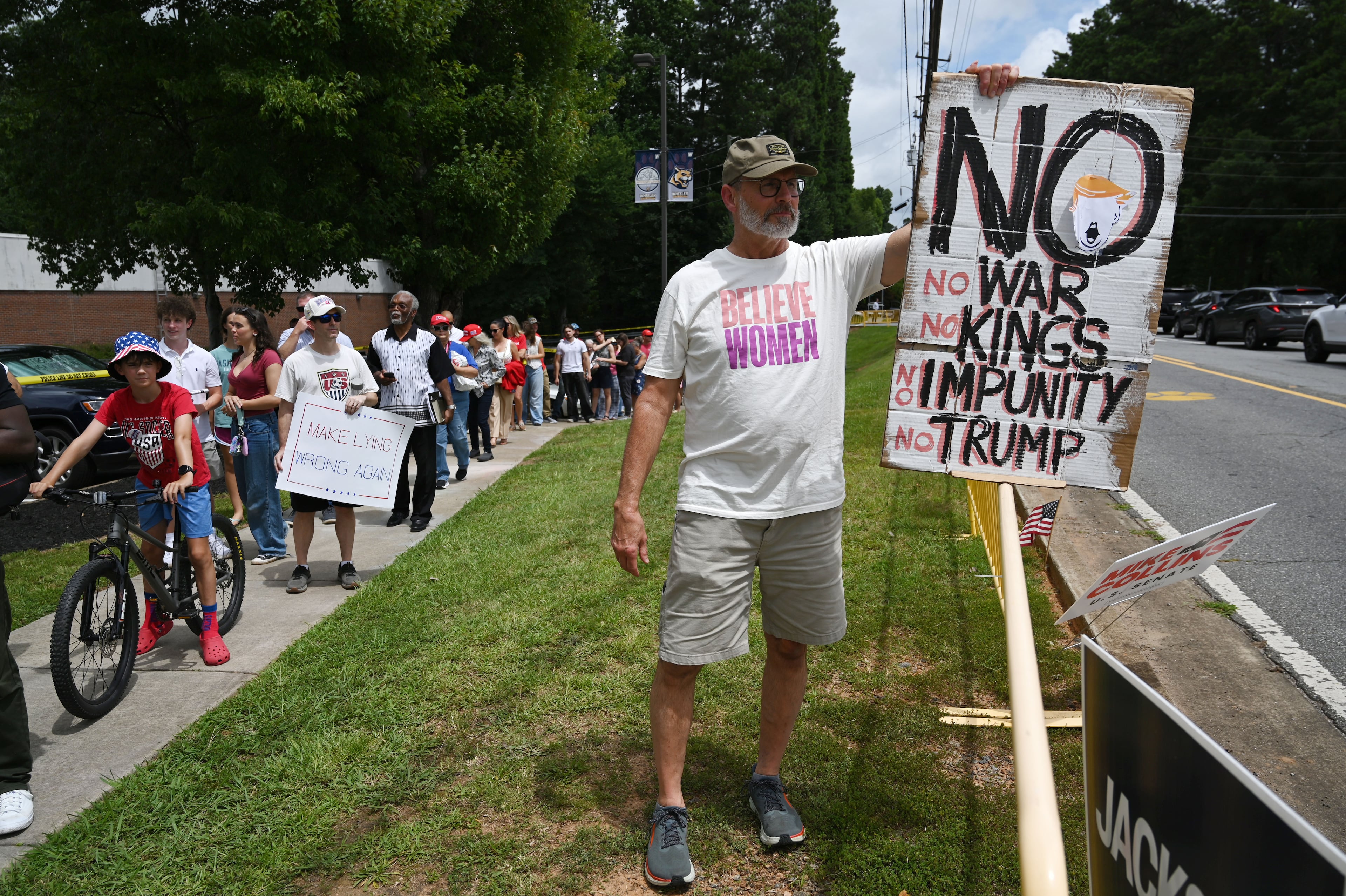 Phil King, right, holds a sign as President Donald Trump supporters, left, arrive outside Wheeler High School on Wednesday, July 22, 2026, in Marietta. President Donald Trump is visiting Cobb County’s Wheeler High School on Wednesday to promote a new investment account for children. (Hyosub Shin/AJC)