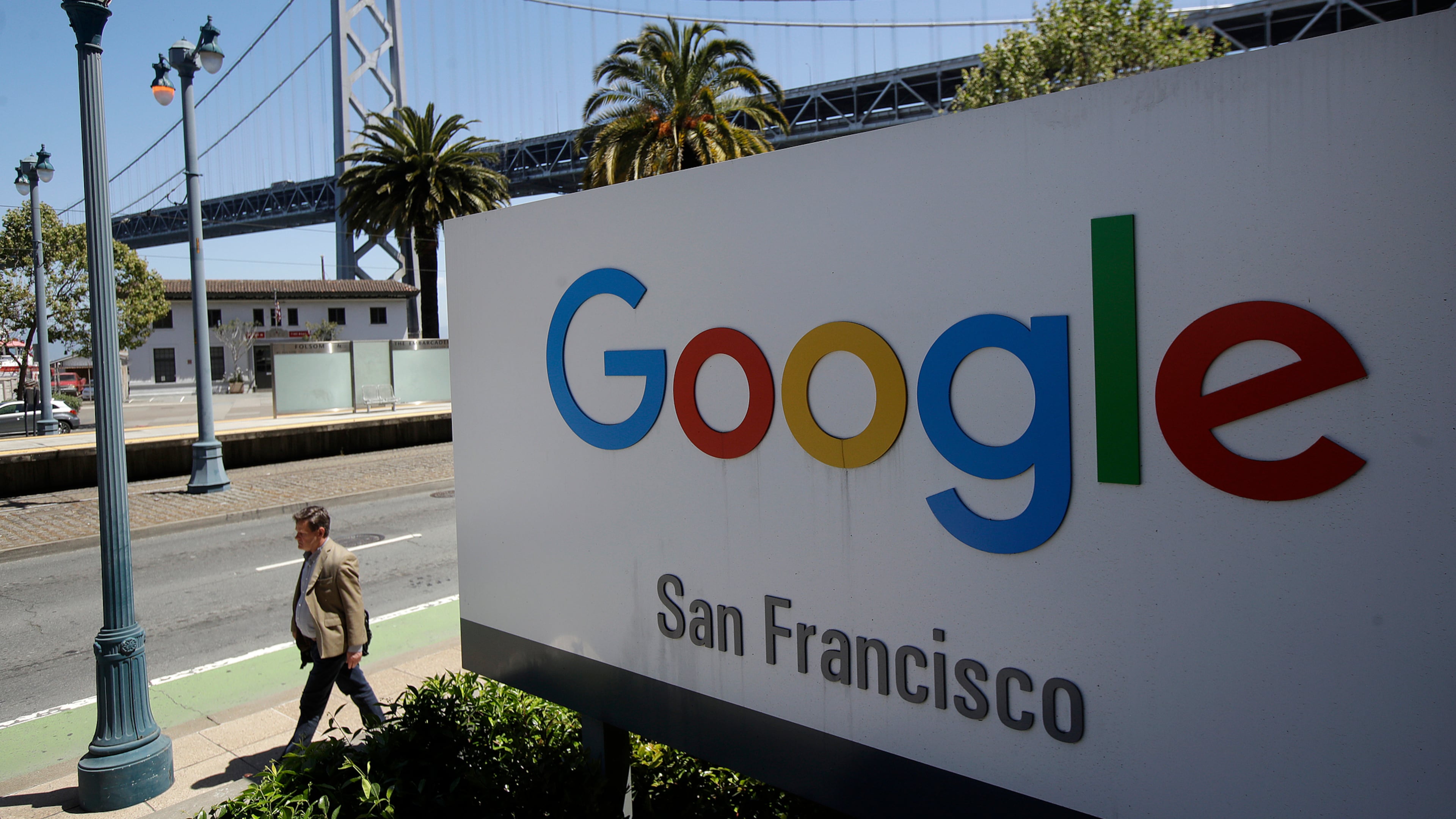 FILE - In this May 1, 2019, file photo a man walks past a Google sign outside with a span of the Bay Bridge at rear in San Francisco. (AP Photo/Jeff Chiu, File)