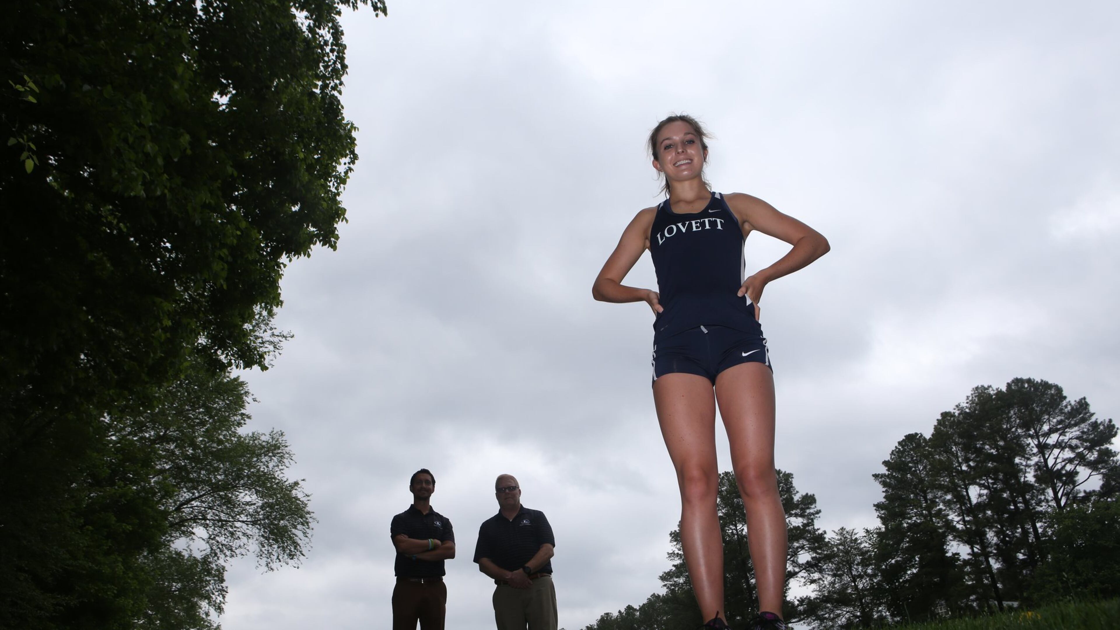 Serena Tripodi (right) poses for a portrait with her coaches Jimmy Jewell (center) and Chris Mayer (left) in the background at The Lovett School in Atlanta, Georgia, on May 4, 2017. Tripodi holds seven state titles in track and cross country, with five of those being individual. (HENRY TAYLOR / HENRY.TAYLOR@AJC.COM)
