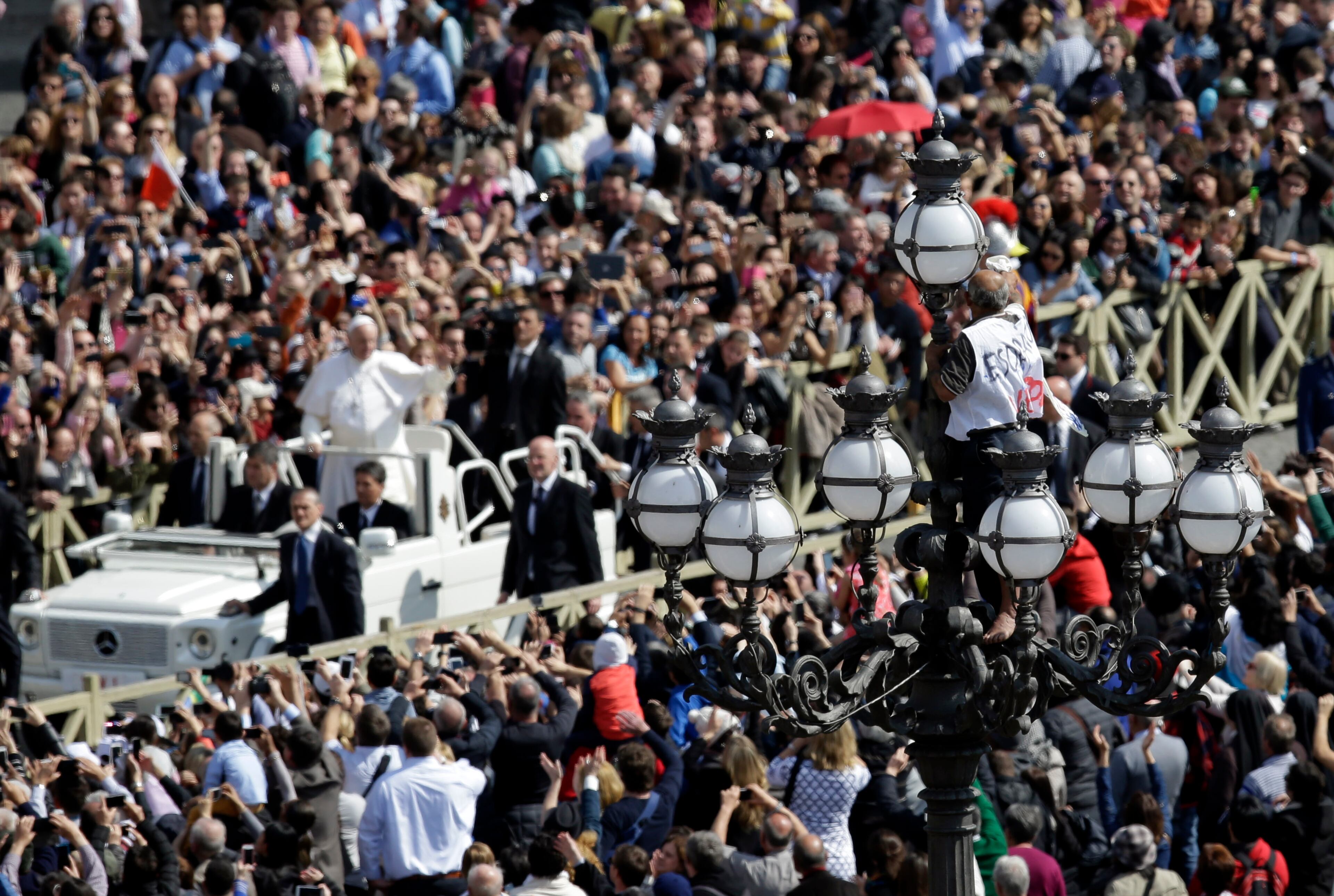 A man climbs on a street lamp as Pope Francis, background, leaves at the end of the Easter Mass, in St. Peter's Square, at the Vatican, on March 27, 2016.