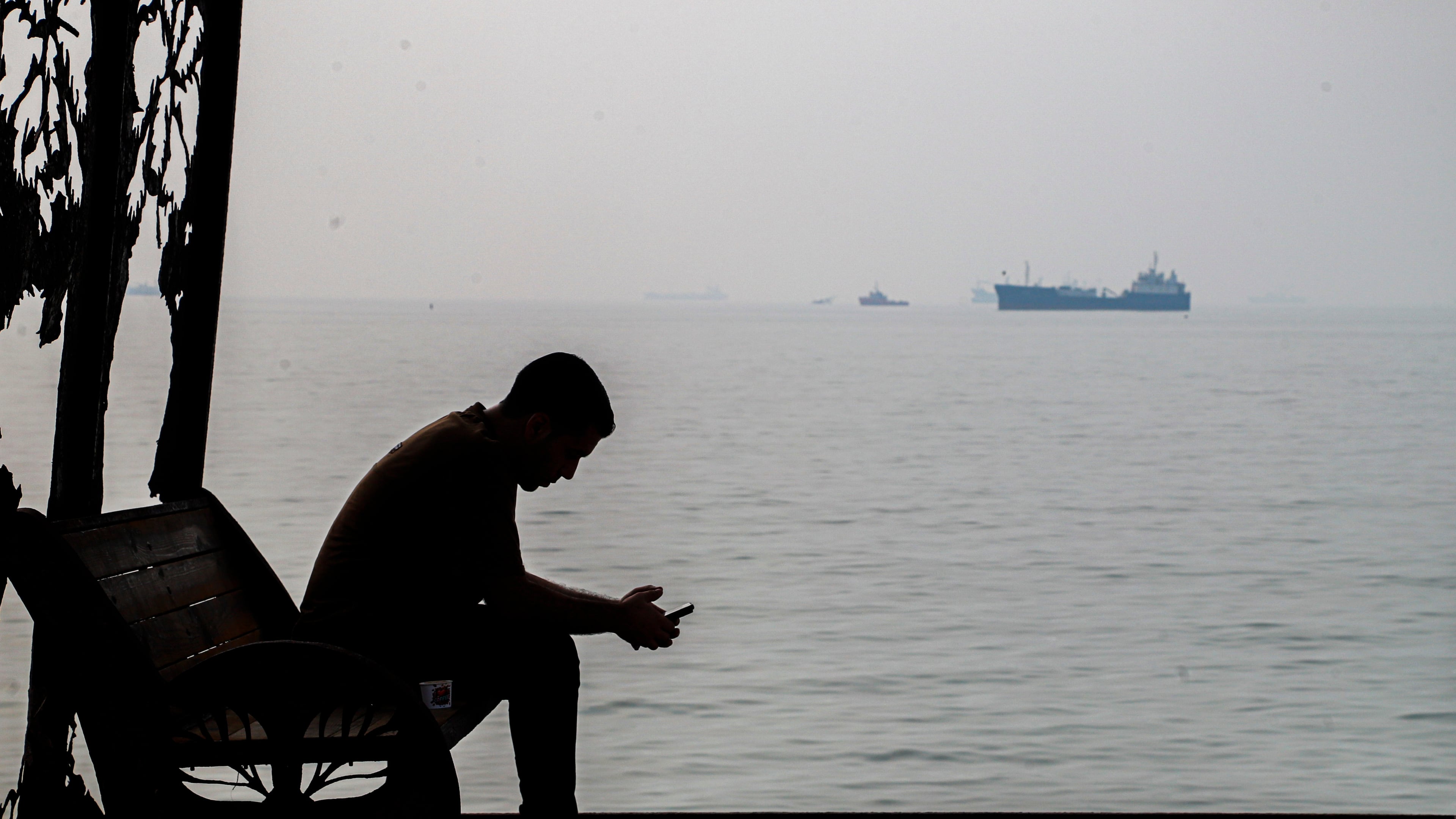 A man checks his cellphone at a waterfront shelter as commercial vessels appear anchored in the Strait of Hormuz off Bandar Abbas, Iran, Monday, Tuesday, Oct, 6, 2026. (Amirhosein Khorgooi/ISNA via AP)