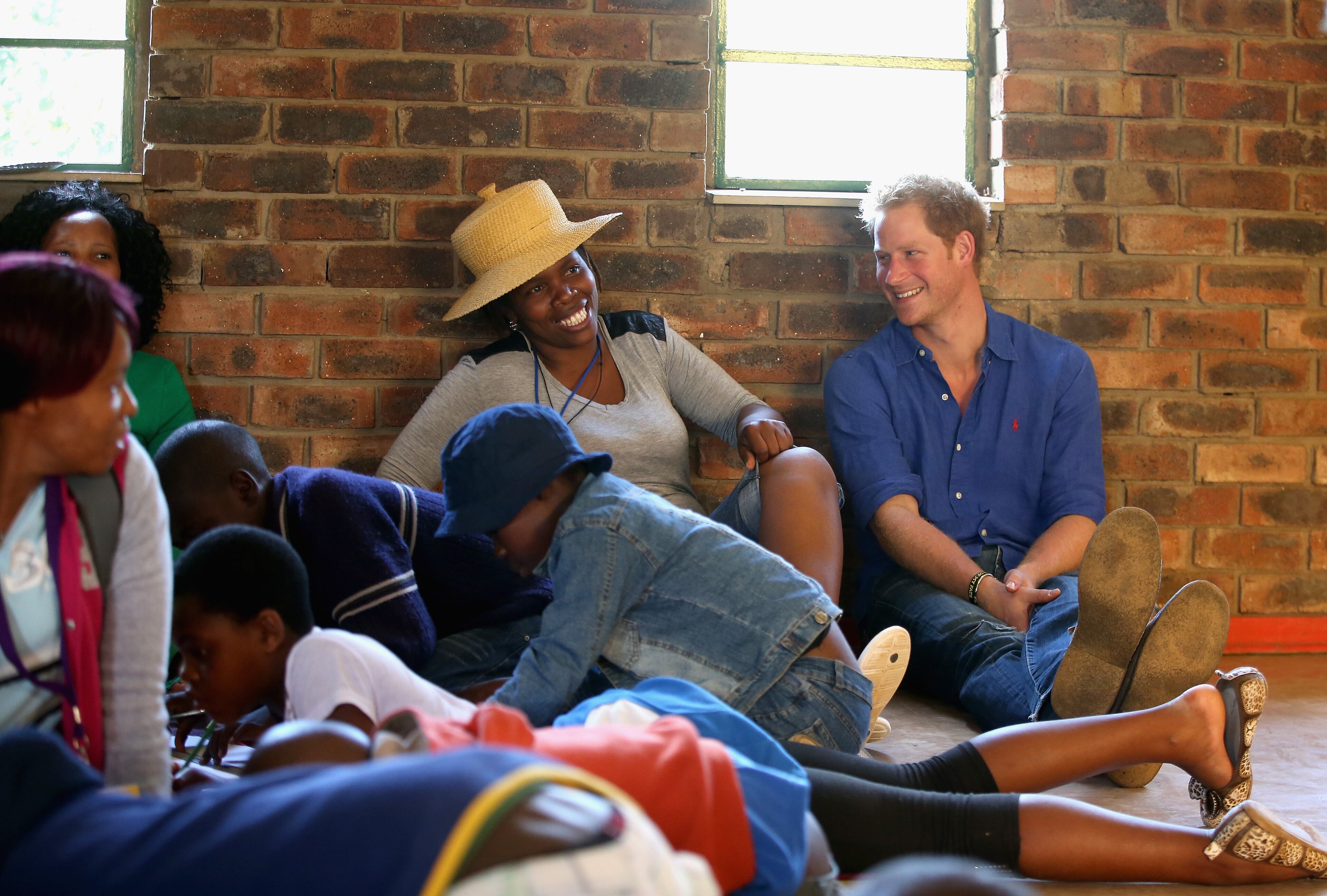 Prince Harry sits next to a volunteer who was laughing at his singing during a visit to a Mamohato Network Club for children living with HIV at St Paul Centre on December 9, 2014 in Maseru, Lesotho. Prince Harry was visiting Lesotho to see the work of his charity Sentebale. Sentebale provides healthcare and education to vulnerable children in Lesotho, Southern Africa. Prince Harry said "A chance to see one of the camps in full swing. Normally when we visit Lesotho, it's school time and therefore we always miss the Camp Mamohato. 44 children, from all over Lesotho gathered for a week of fun and learning about living with HIV. These are children who have never had the chance to talk about their illness, and who had no idea that they were one of so many in their age group. It was really emotional watching them interact with each other. Some really outgoing chatty kids, others slightly overwhelmed, but all with huge smiles. This confirmed to me again that what we're doing is going to change thousands of children's lives, and hopefully save a generation." (Photo by Chris Jackson - WPA Pool /Getty Images)