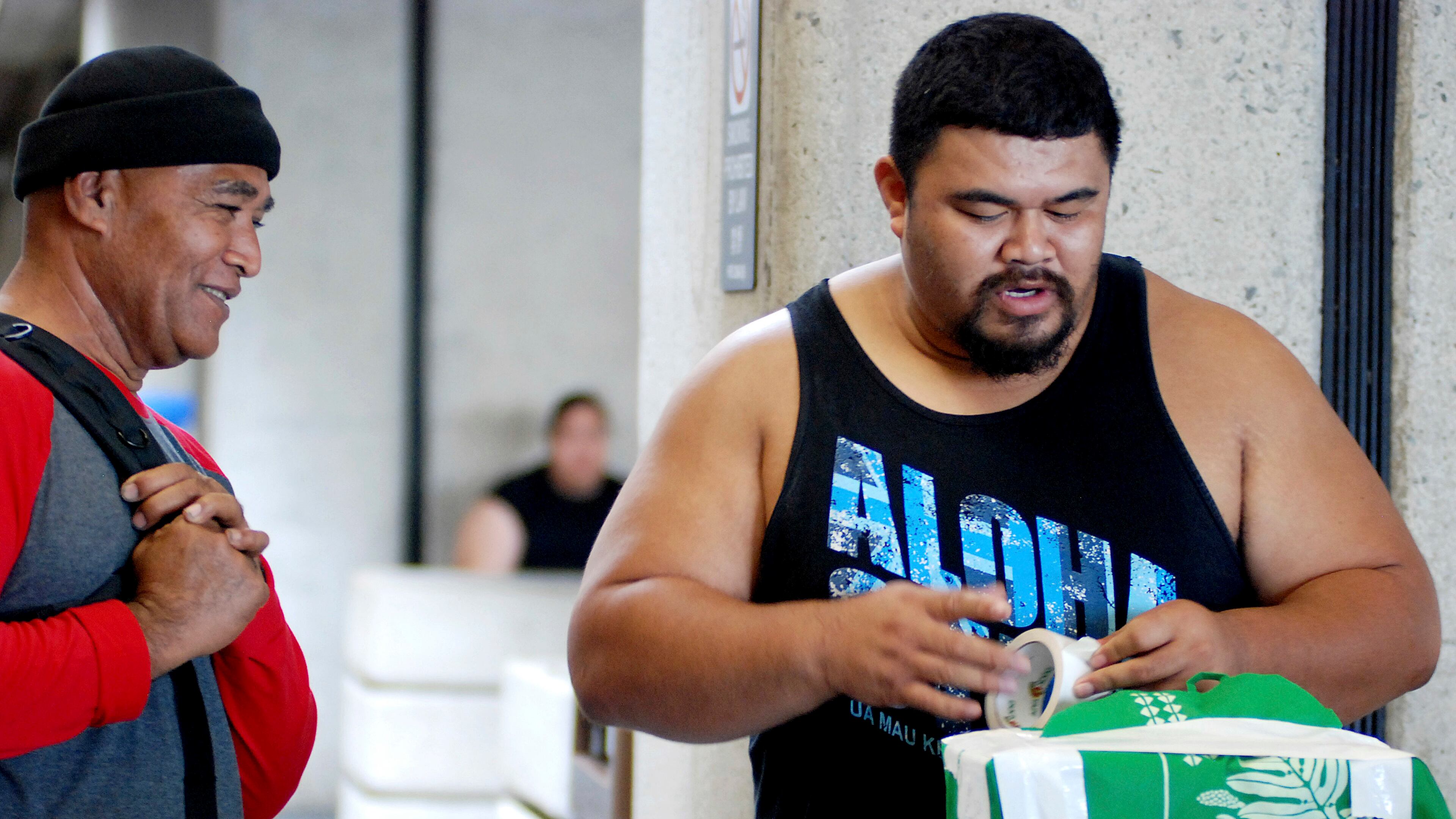 In this Oct. 10, 2016 photo, Mua Migi, left, watches his son Atimua Migi put tape on a carry-on bag before boarding a Hawaiian Airlines flight to Pago Pago, American Samoa at Honolulu International Airport in Honolulu. Atimua Migi doesn't like the airline's new policy of assigning seats at the airport after the airline conducted a survey that involved weighing passengers traveling between Pago Pago and Honolulu. (AP Photo/Jennifer Sinco Kelleher)