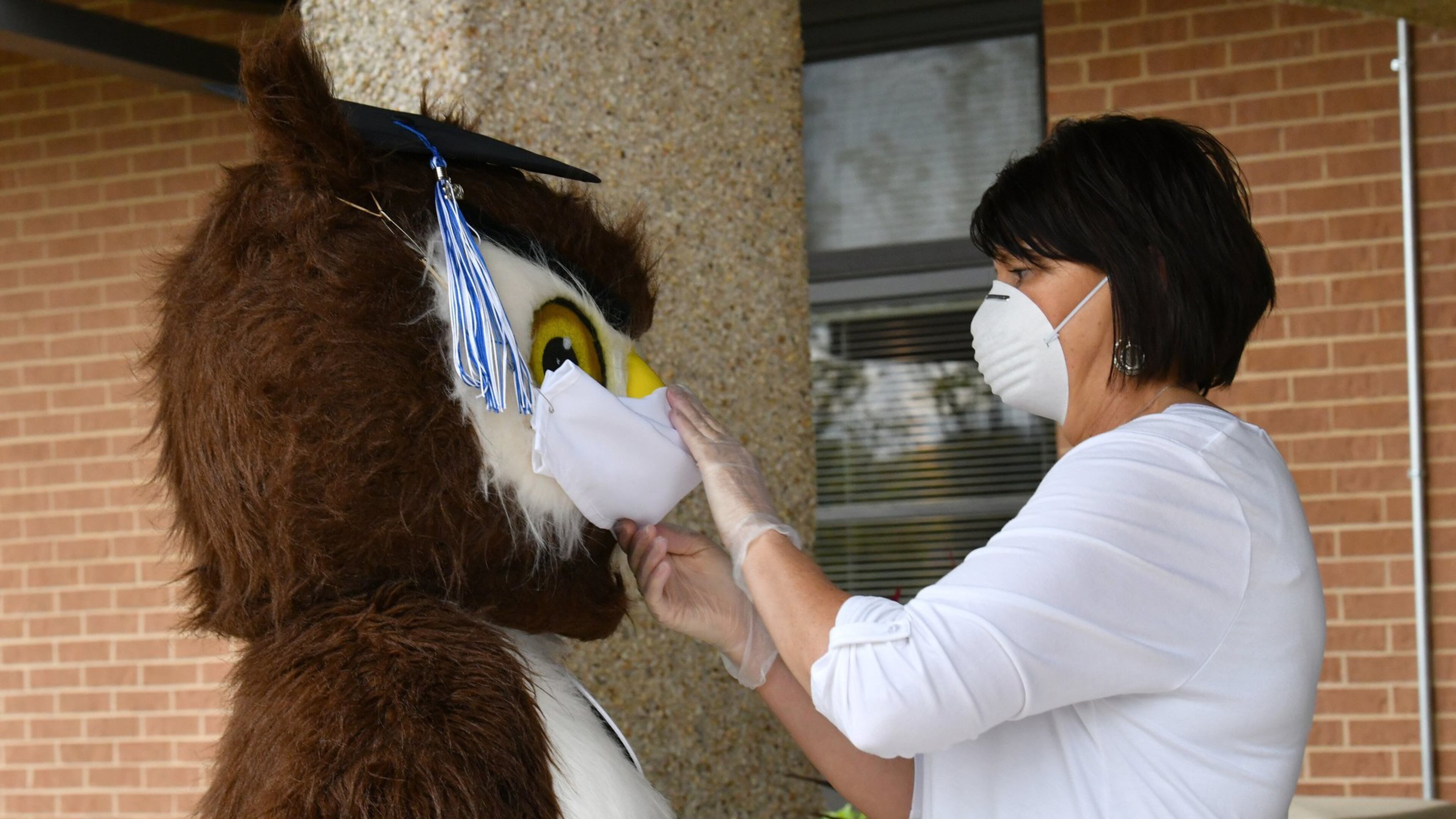 Petra Ordini (right), with office of student services, adjusts a face mask of Gwinnett Online Campus mascot Dr. Whooo, performed by Shannon Williams, as they wait for students picking up their caps and gowns last year outside the main door of Gwinnett Online Campus in Lawrenceville in 2020 .Hyosub Shin/Hyosub.Shin@ajc.com