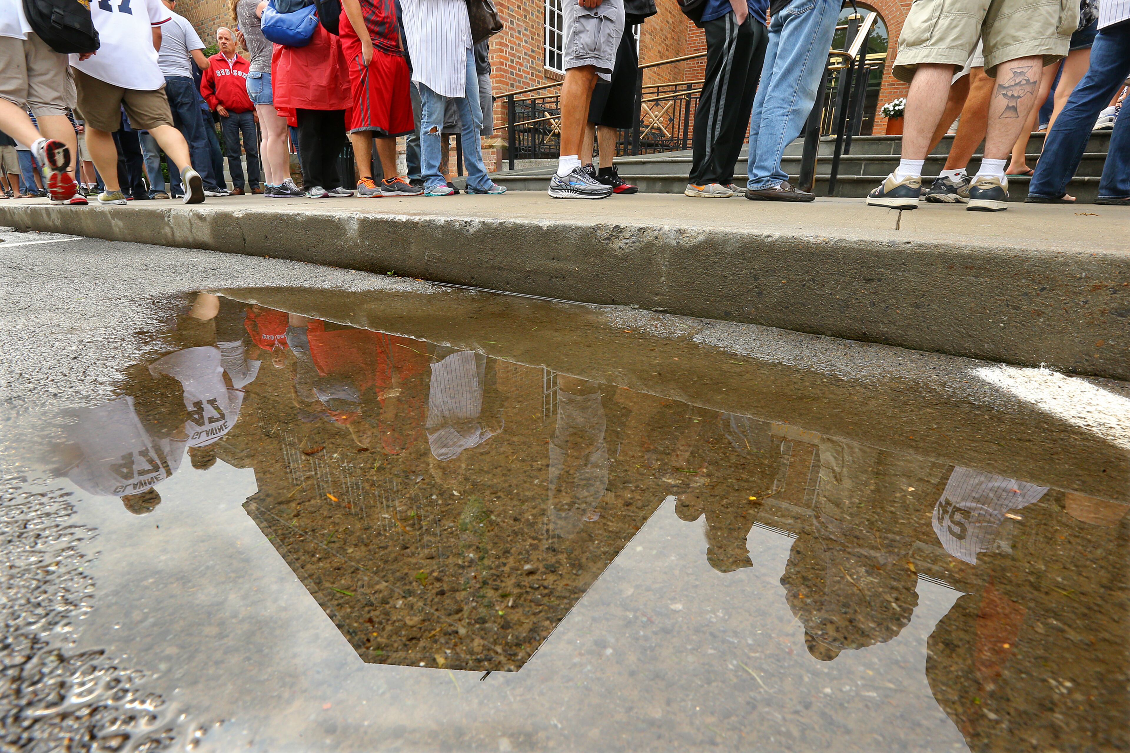 Hundreds of baseball fans line up upside the National Baseball Hall of Fame despite the weather and are reflected in a puddle of rain water along with the building on Sunday, July 27, 2014, in Cooperstown. The annual Hall of Fame Induction Ceremony will take place nearby at the Clark Sports Center.