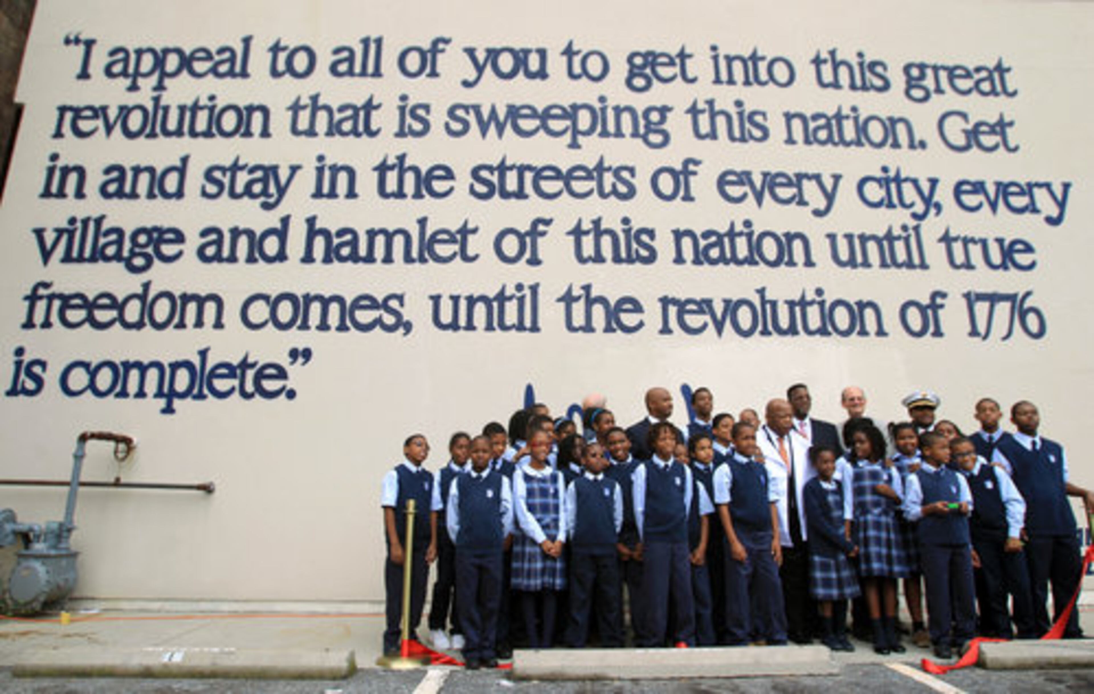 Lewis (center in white coat), poses for a photograph with officials and students from Imhotep Academy in front of the mural. On this wall is written a portion of Lewis' speech from the March on Washington on August 28, 1963.
