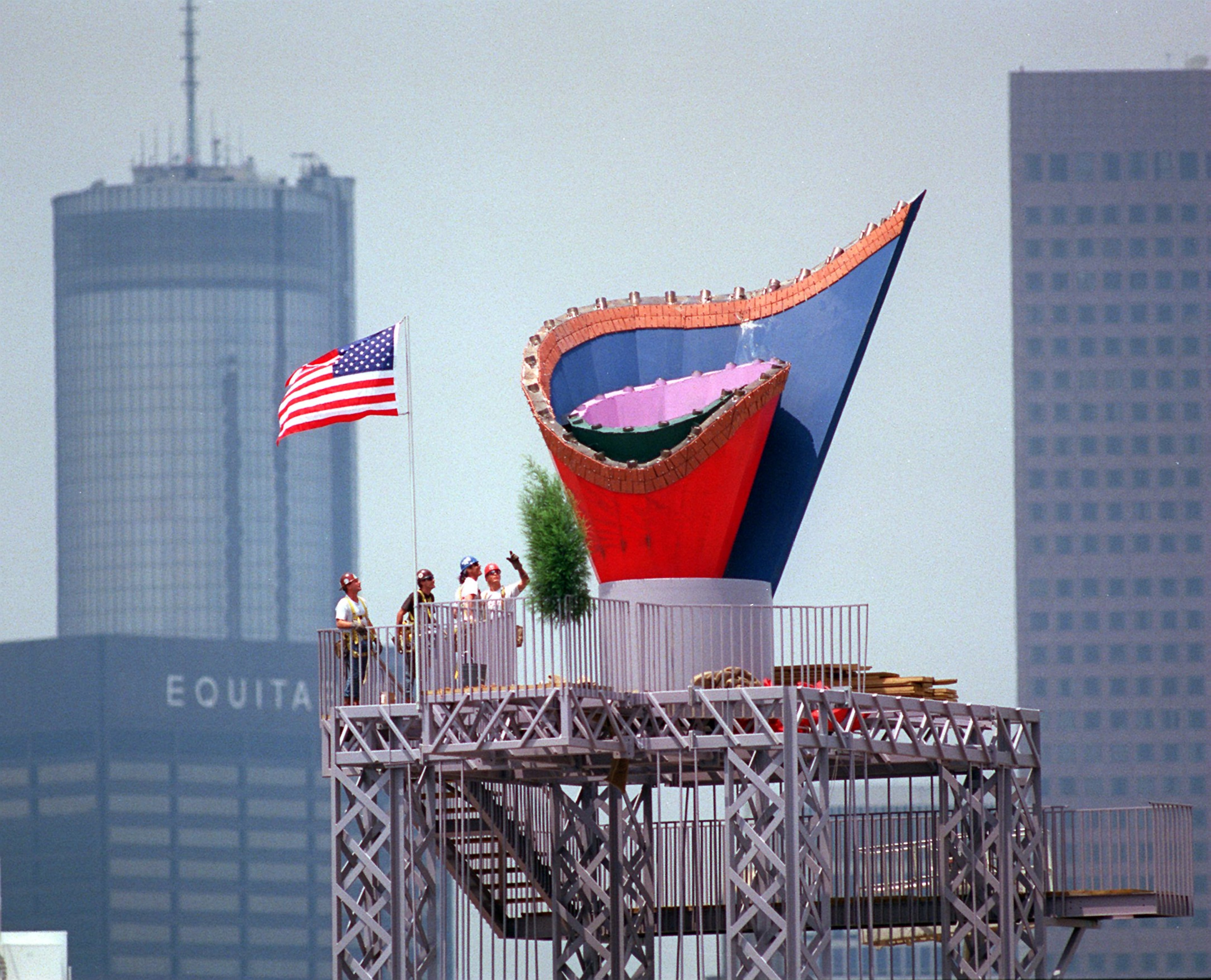 With a flag completing the picture, ironworkers admire their efforts after installing the Olympic cauldron atop its 116-foot-high tower on Thursday, May 30, 1996, ahead of the Olympic Games in Atlanta. The structure consisted of three parts: the cauldron, the cauldron tower and a bridge that connected it to the stadium. The cauldron, made of stainless steel, has a terra cotta rim of Georgia red clay. (Joey Ivansco/AJC)