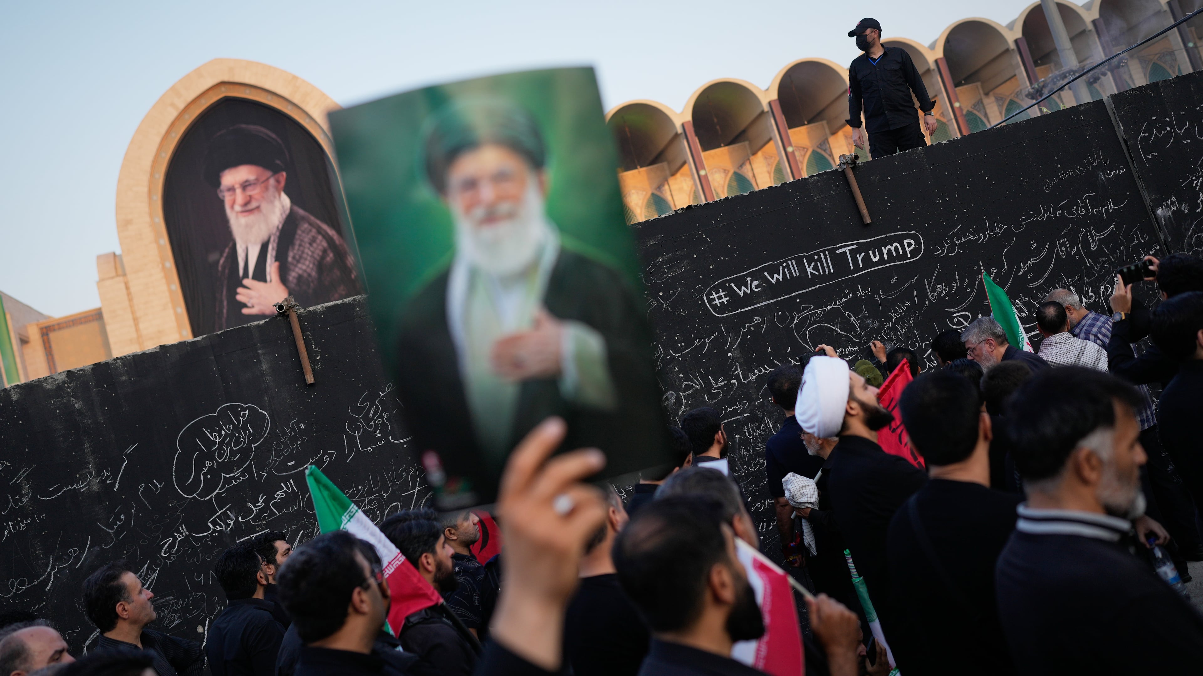 A mourner holds a portrait of the late Iranian Supreme Leader Ayatollah Ali Khamenei as he walks past a wall bearing messages, including one in English that reads "We will kill Trump," during the funeral ceremonies for Khamenei and members of his family at the Imam Khomeini Mosalla Grand Mosque in Tehran, Iran, Saturday, July 4, 2026. (AP Photo/Altaf Qadri)