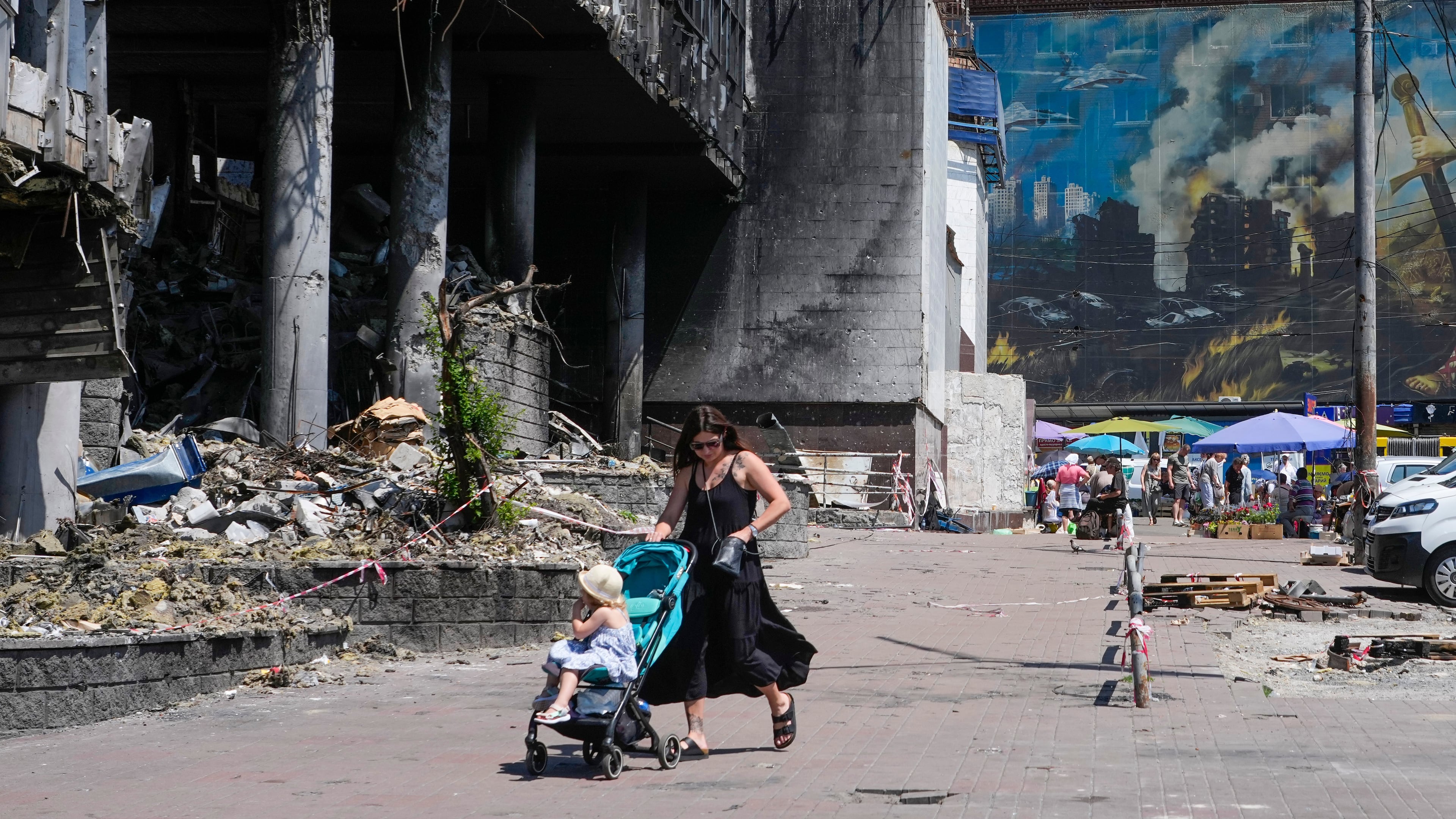 A mother pushes a stroller past a damaged building covered with street artist paintings and a big city marketplace that was destroyed recently by Russian missiles in Kyiv, Ukraine, Monday, June 22, 2026. (AP Photo/Efrem Lukatsky)