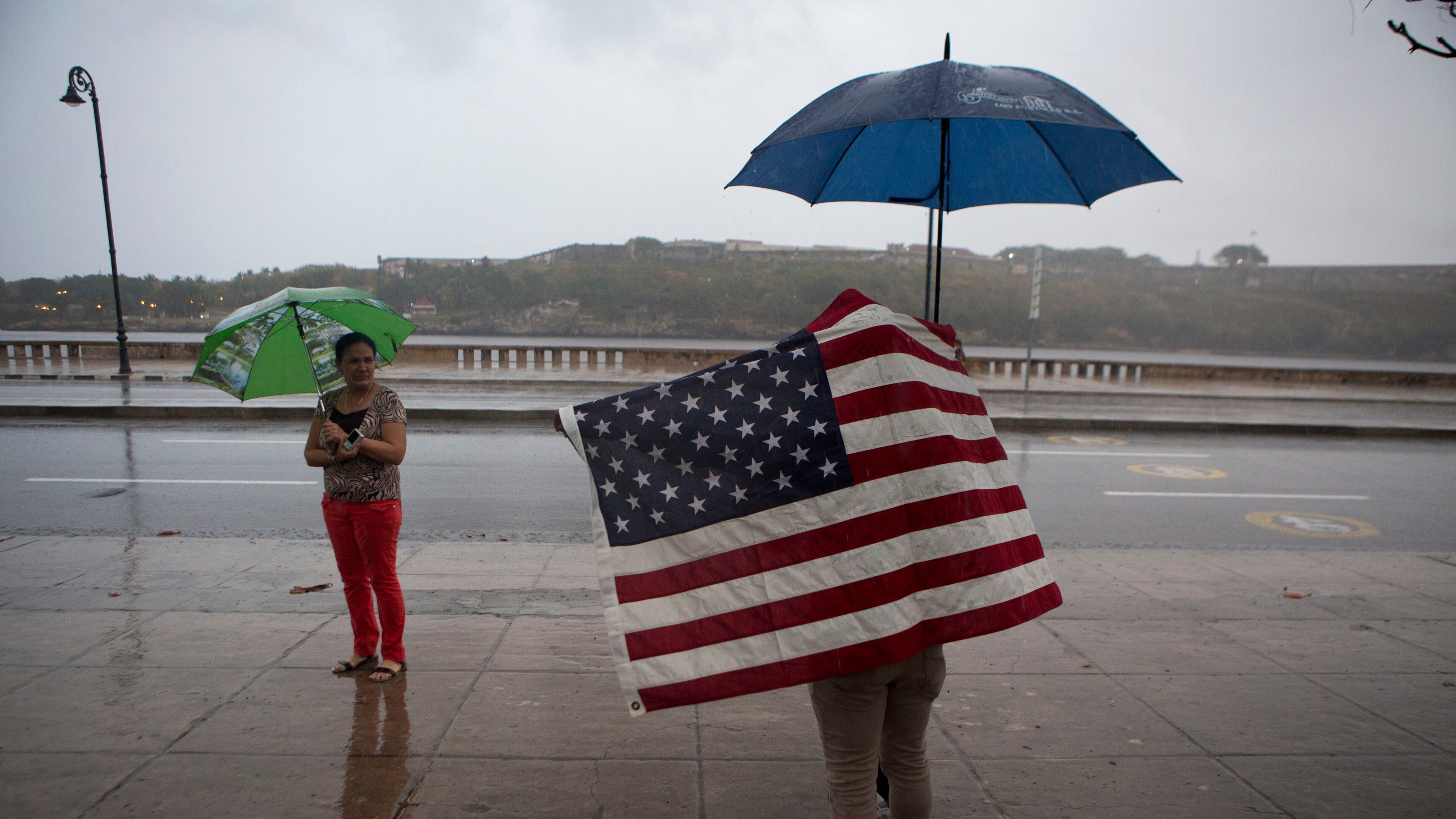 A Cuban man carrying the Stars and Stripes waits in the rain to wave to U.S. President Barack Obama's convoy as it arrives along the Malecon into Old Havana, Cuba, Sunday, March 20, 2016. Obama's trip is a crowning moment in his and Cuban President Raul Castro's ambitious effort to restore normal relations between their countries. (AP Photo/Rebecca Blackwell)