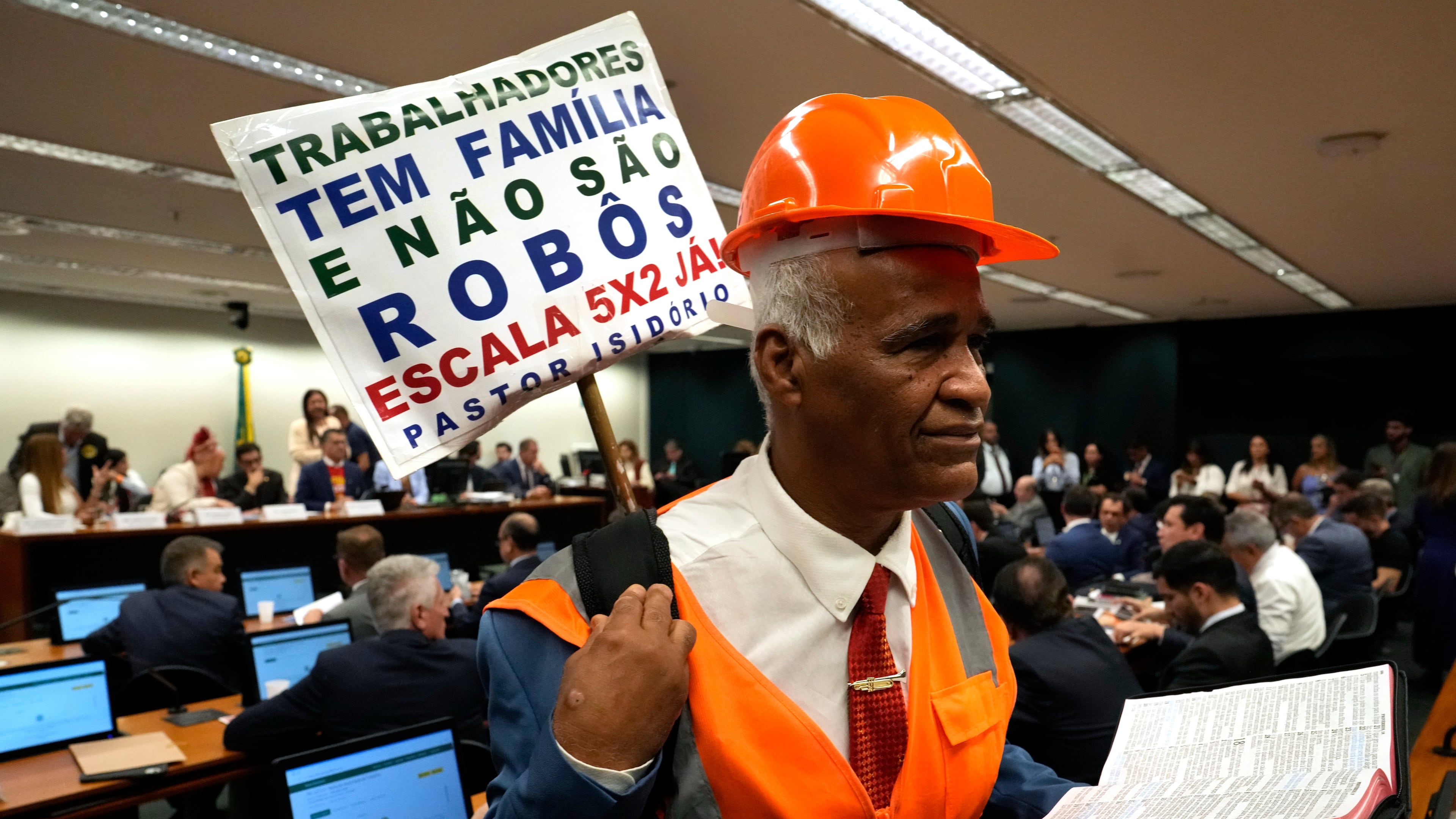 Lamaker and Pastor Sargento Isidorio holds a sign supporting the end of Brazil's six-day workweek schedule, that reads in Portuguese: "Workers have families and are not robots. 5x2 schedule now," during a special committee session analyzing the proposal at the Chamber of Deputies in Brasilia, Brazil, Wednesday, May 27, 2026. (AP Photo/Eraldo Peres)