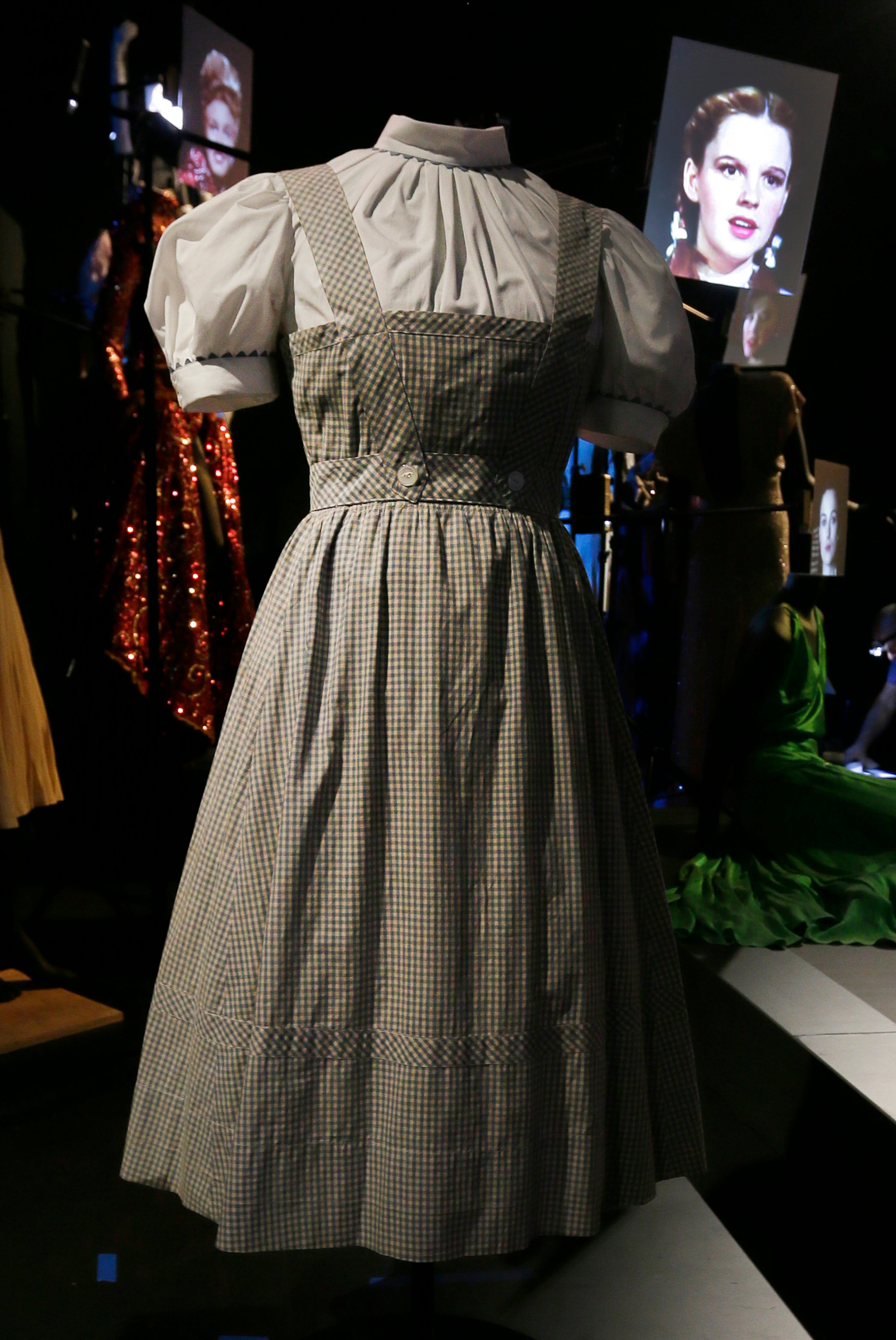 The blue and white gingham pinafore dress worn by Judy Garland in her iconic role of Dorothy designed by Adrian for the 1939 film The Wizard Of Oz, with a video portrait of garland in the background.