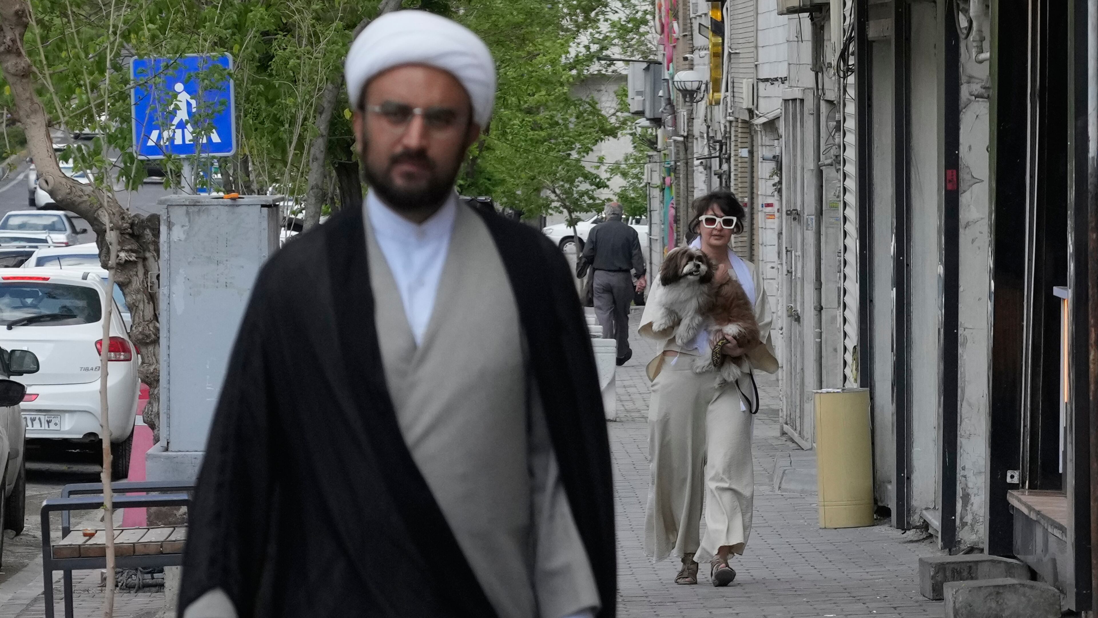 A woman carries her dog as a cleric walks on a sidewalk in Tehran, Iran, Tuesday, April 21, 2026. (AP Photo/Vahid Salemi)