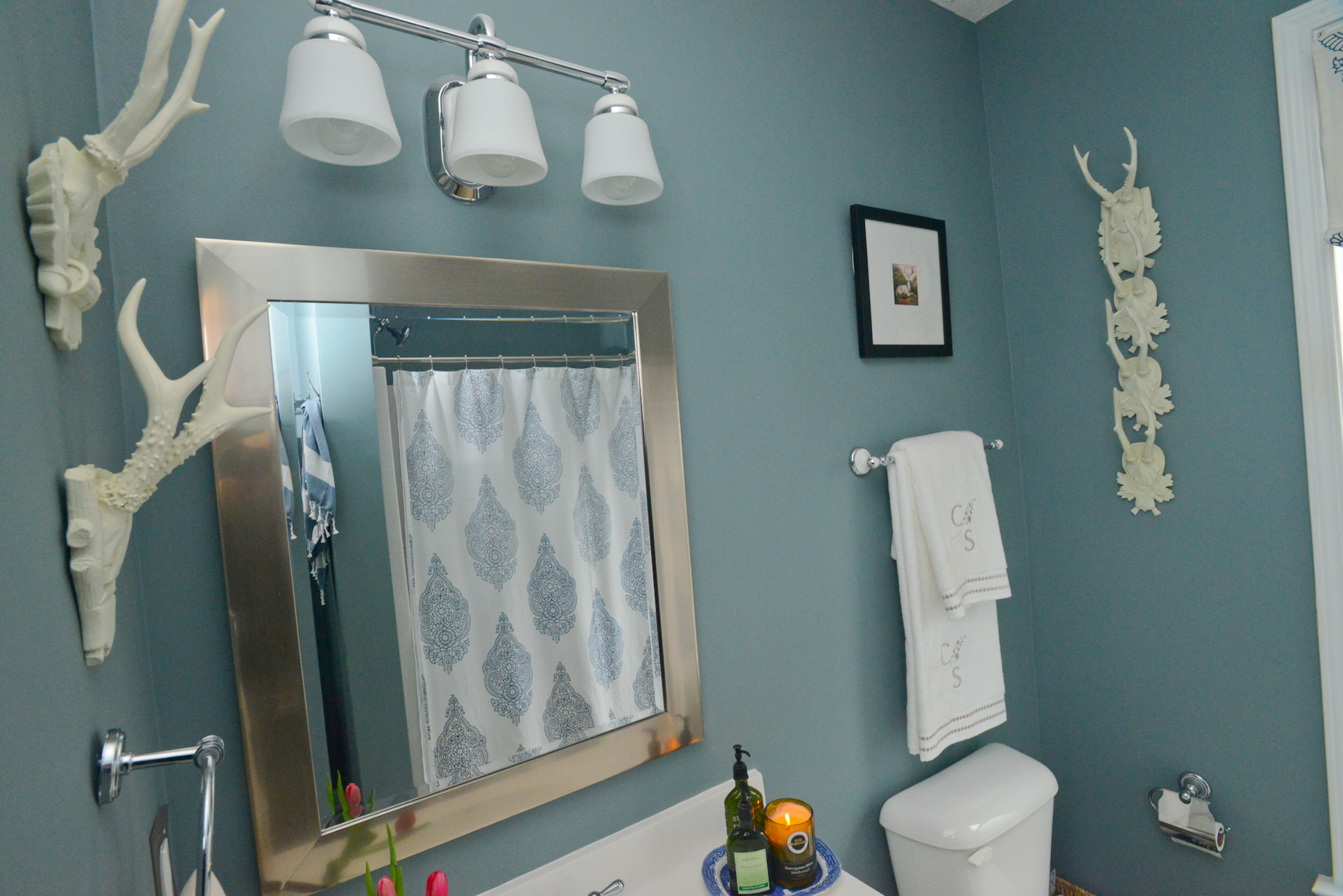 A soft blue backdrop highlights white lighting fixtures and antler wall accessories from Ballard Designs in the guest bathroom. The shower curtain and towels are from Pottery Barn.