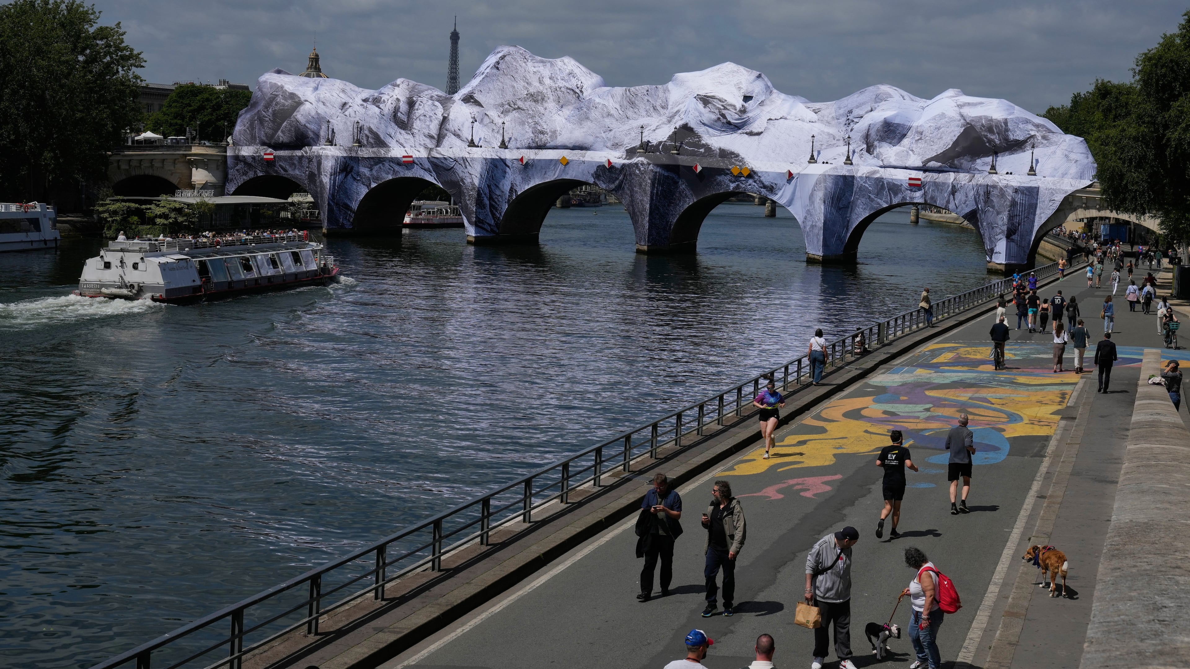 People walk along the Seine river next to "The Pont Neuf Cave," an inflated art installation by French street artist JR, on Paris' oldest bridge, the Pont Neuf, Thursday, May 21, 2026, which will be open to the public from June 6-28. (AP Photo/Michel Euler)