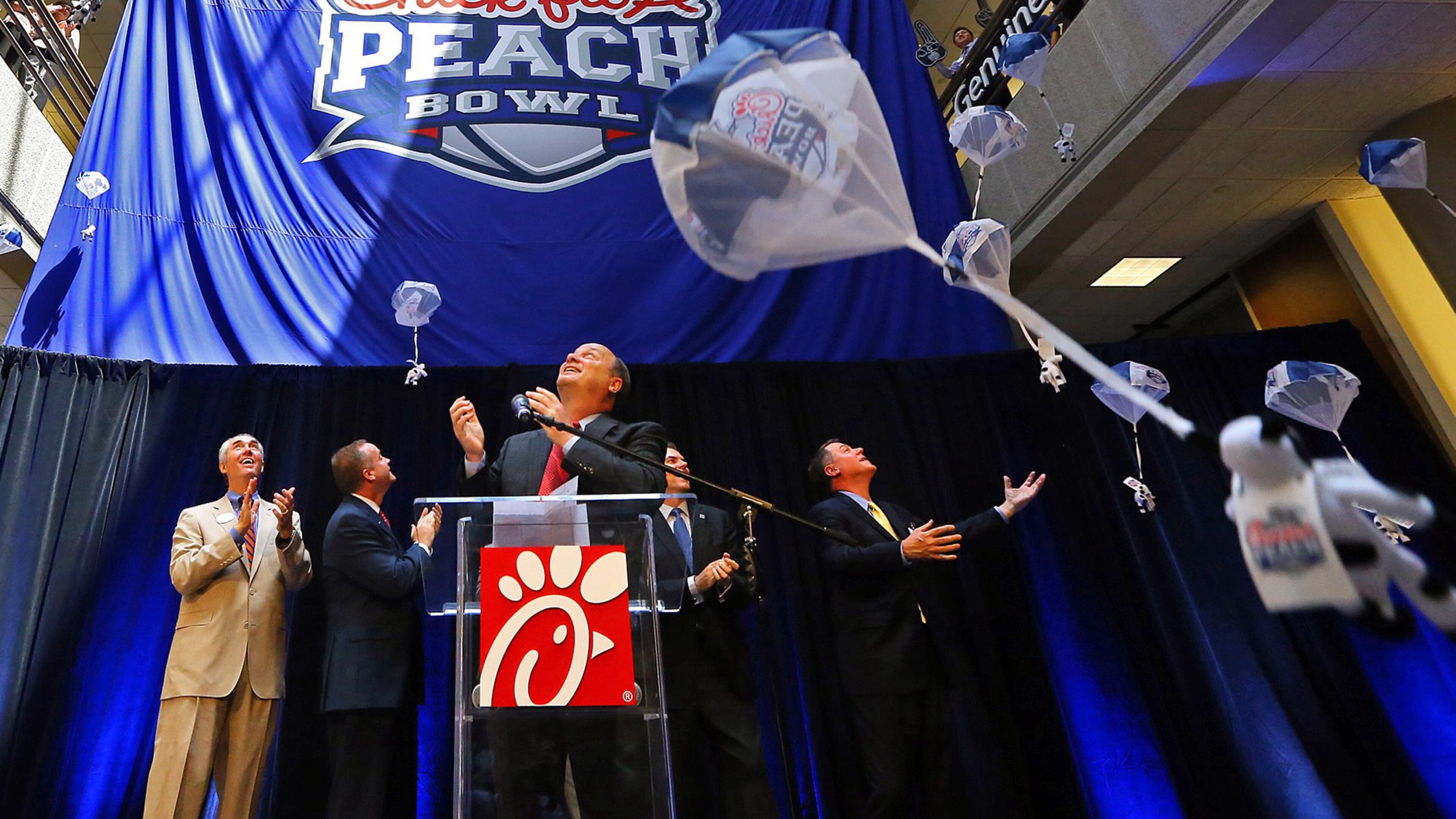 Steve Robinson (from left), Chick-fil-A executive vice president; Michael Kelly, College Football Playoff executive; Gary Stokan, bowl president; Rob Temple, ESPN vice president; and Burke Magnus, ESPN senior vice president, look up as the bowl’s new name Chick-fil-A Peach Bowl logo is unveiled while parachuting cows drop from the ceiling Monday, April 21, 2014, in Atlanta. CURTIS COMPTON / AJC file