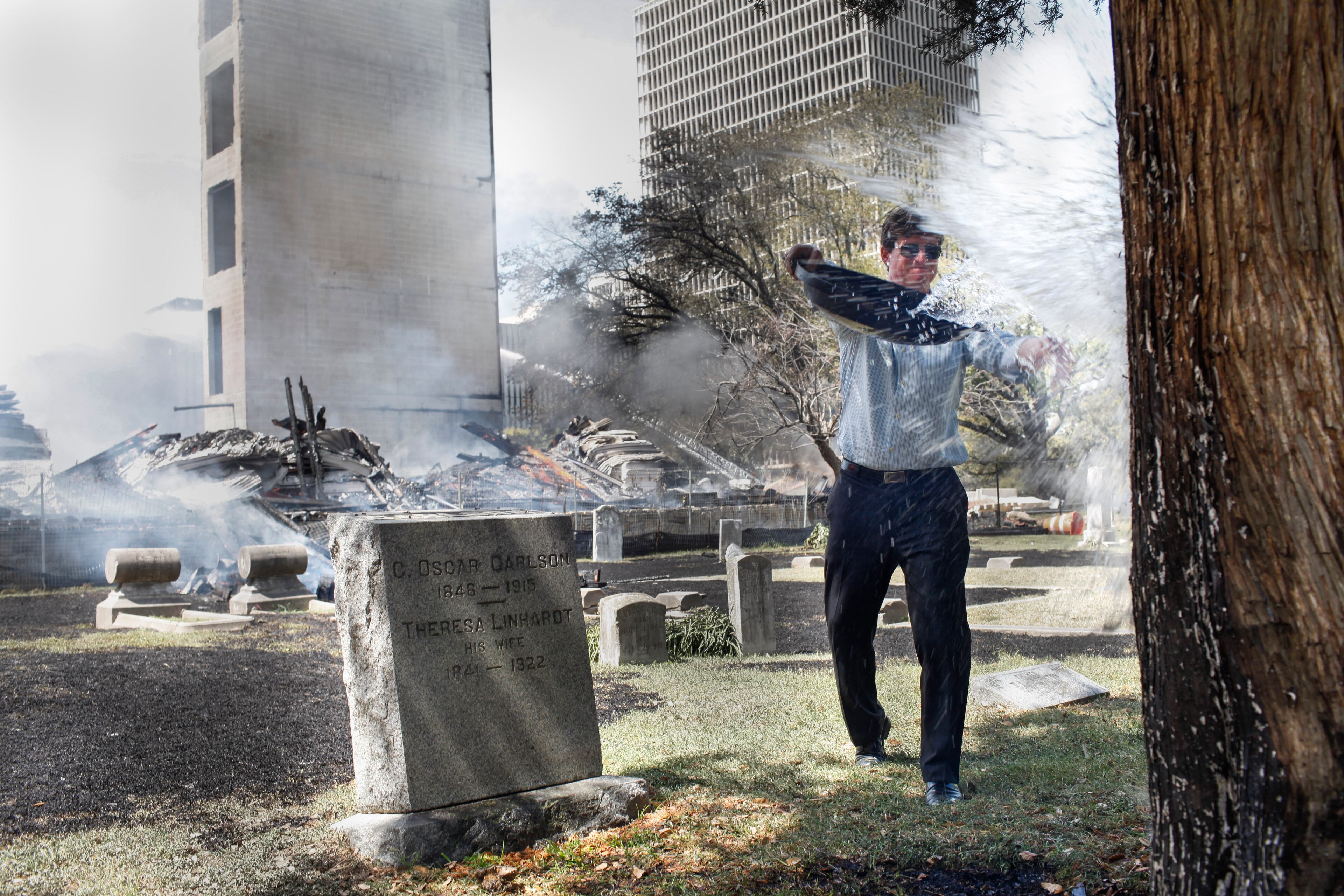 Concerned citizen Craig Guidry splashes water on a smoldering cypress tree in Magnolia Cemetery as five-alarm fire burn at a nearby construction site Tuesday, March 25, 2014, in Houston. Fire officials said more than 200 emergency personnel were at the scene Tuesday afternoon and were working to protect nearby buildings. (AP Photo/Houston Chronicle, Eric Kayne)