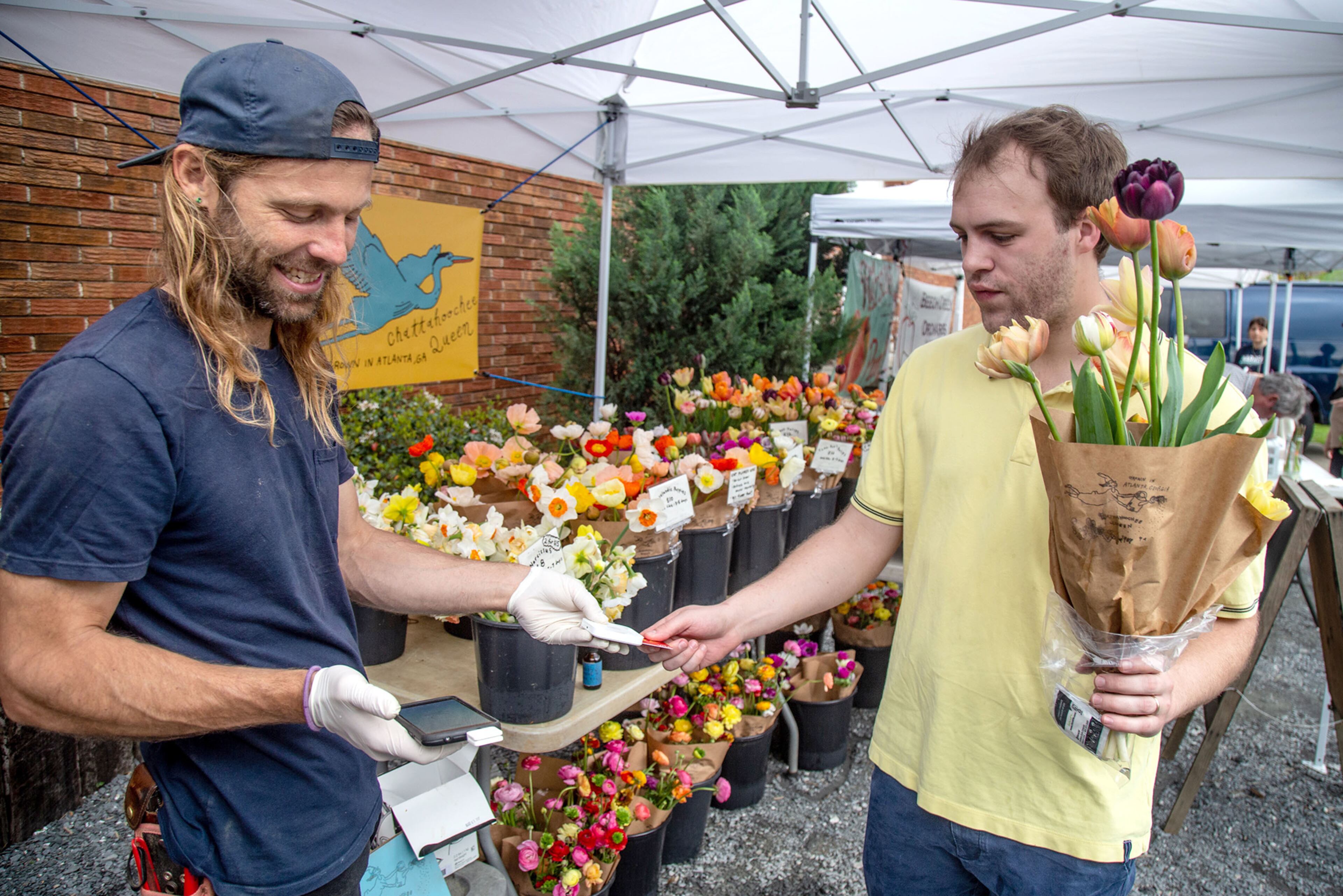 Evan Neal (L) takes a payment for flowers from Walker Loucks at the Grant Park Farmers Market Sunday, March 29, 2020. STEVE SCHAEFER / SPECIAL TO THE AJC