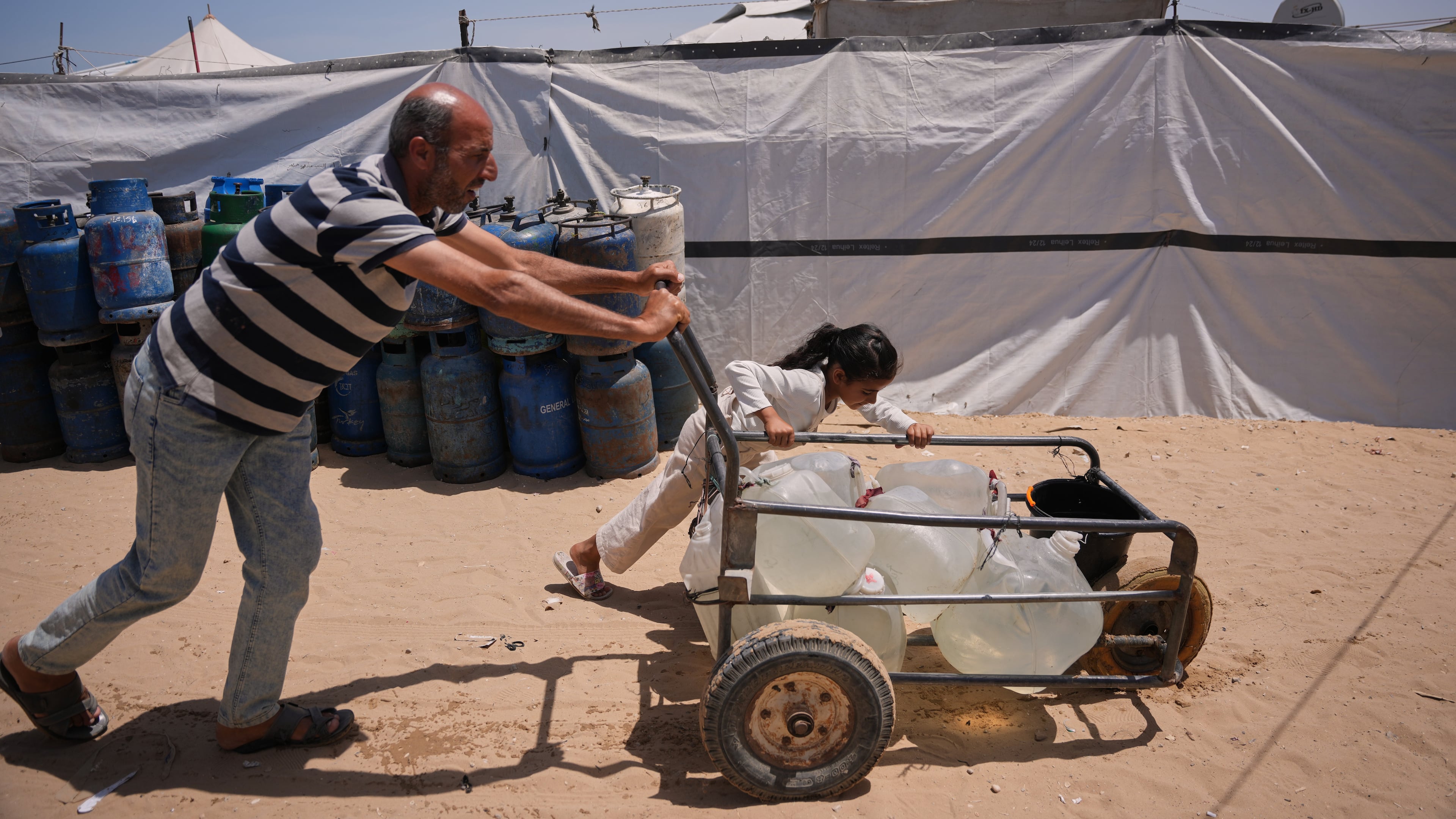 Ne'man Abu Jarad and his daughter push a cart loaded with jerrycans filled with water as they walk at a tent camp for displaced Palestinians in Khan Younis, southern Gaza Strip, Wednesday, May 13, 2026. (AP Photo/Abdel Kareem Hana)