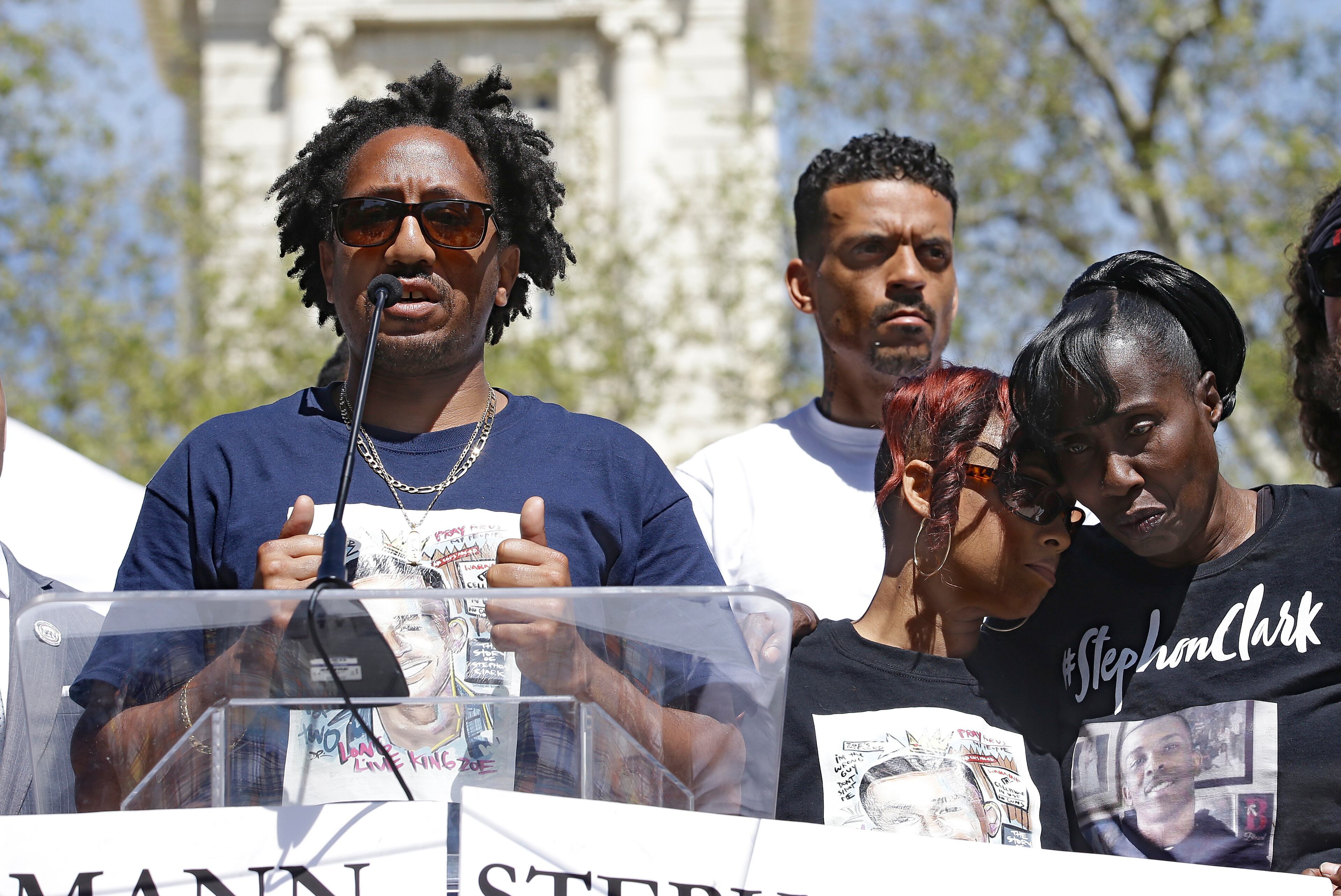 Curtis Gordon, left, the uncle of police shooting victim Stephon Clark, speaks at a rally aimed at ensuring Clark's memory and calling for police reform, Saturday, March 31, 2018, in Sacramento, Calif. The gathering comes nearly two weeks after Clark, who was unarmed, was shot and killed by two Sacramento police officers, in the backyard of his grandmother, Sequita Thompson, right, who is being hugged by Clark's aunt Jamilia Land. Standing in the background is former NBA player Matt Barnes, a Sacramento native, who organized the event and announced he was starting a college scholarship fund for Clark's two sons and children of parents killed by police nationwide. (AP Photo/Rich Pedroncelli)
