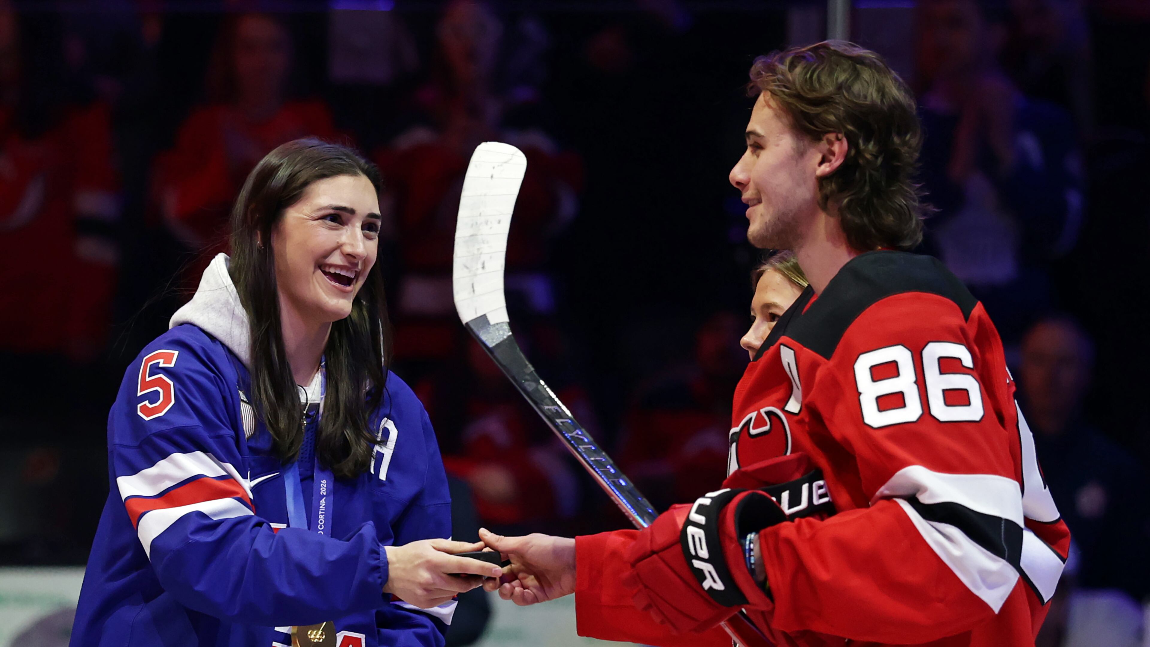 New Jersey Devils center Jack Hughes (86) gives United States women's gold medal hockey players Megan Keller the puck after a ceremonial puck drop before an NHL hockey game Wednesday, March 4, 2026, in Newark, N.J. (AP Photo/Adam Hunger)