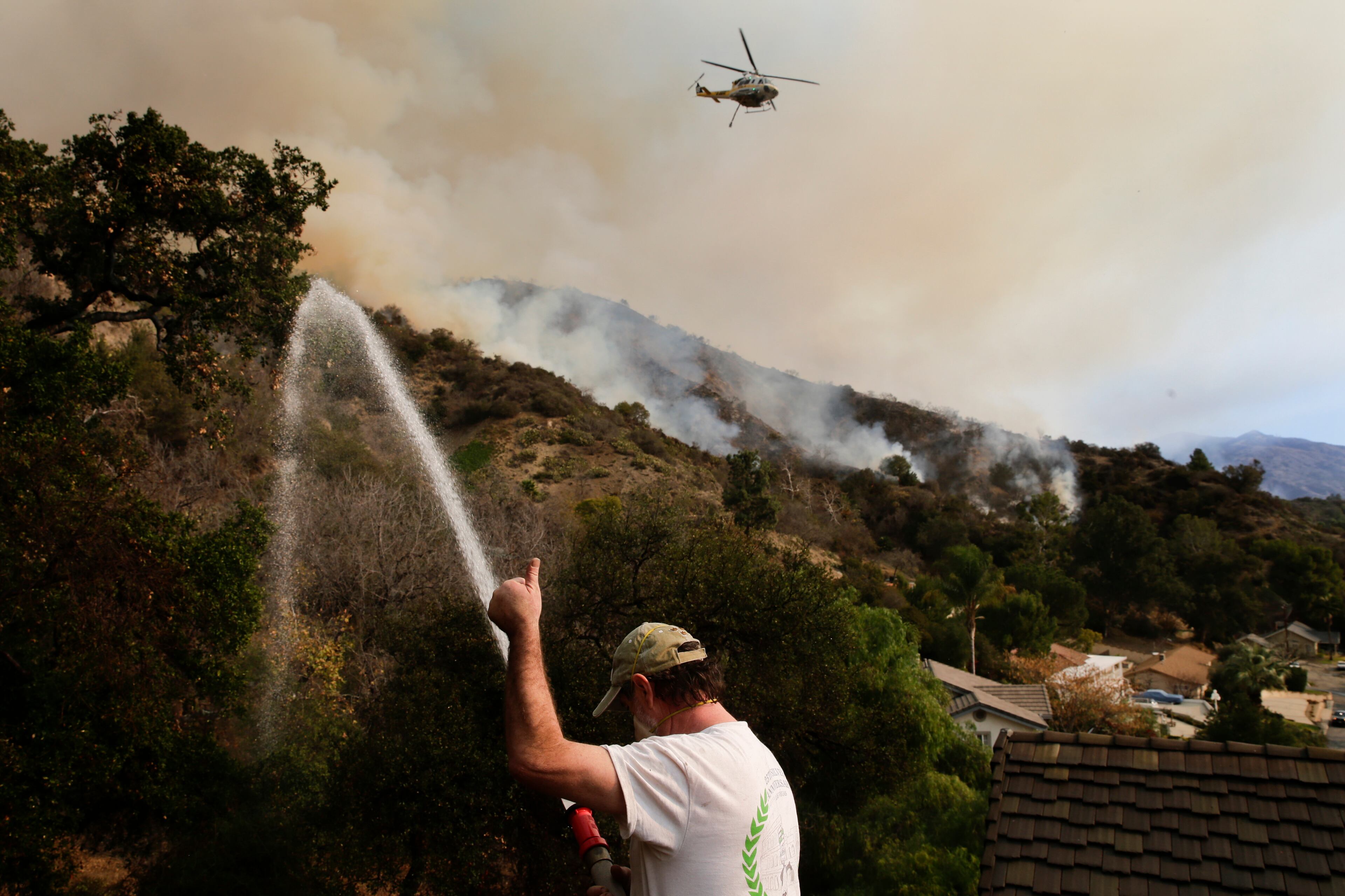 Mark Davis gives a thumbs-up toward a helicopter as he sprays water around his property, Thursday, Jan. 16, 2014, in Azusa, Calif. A wildfire burned out of control near homes in the dangerously dry foothills of the San Gabriel Mountains early Thursday, fanned by gusty Santa Ana winds that spit embers into neighborhoods in the city below, igniting trees. Evacuations were ordered for houses at the edge of the fire.