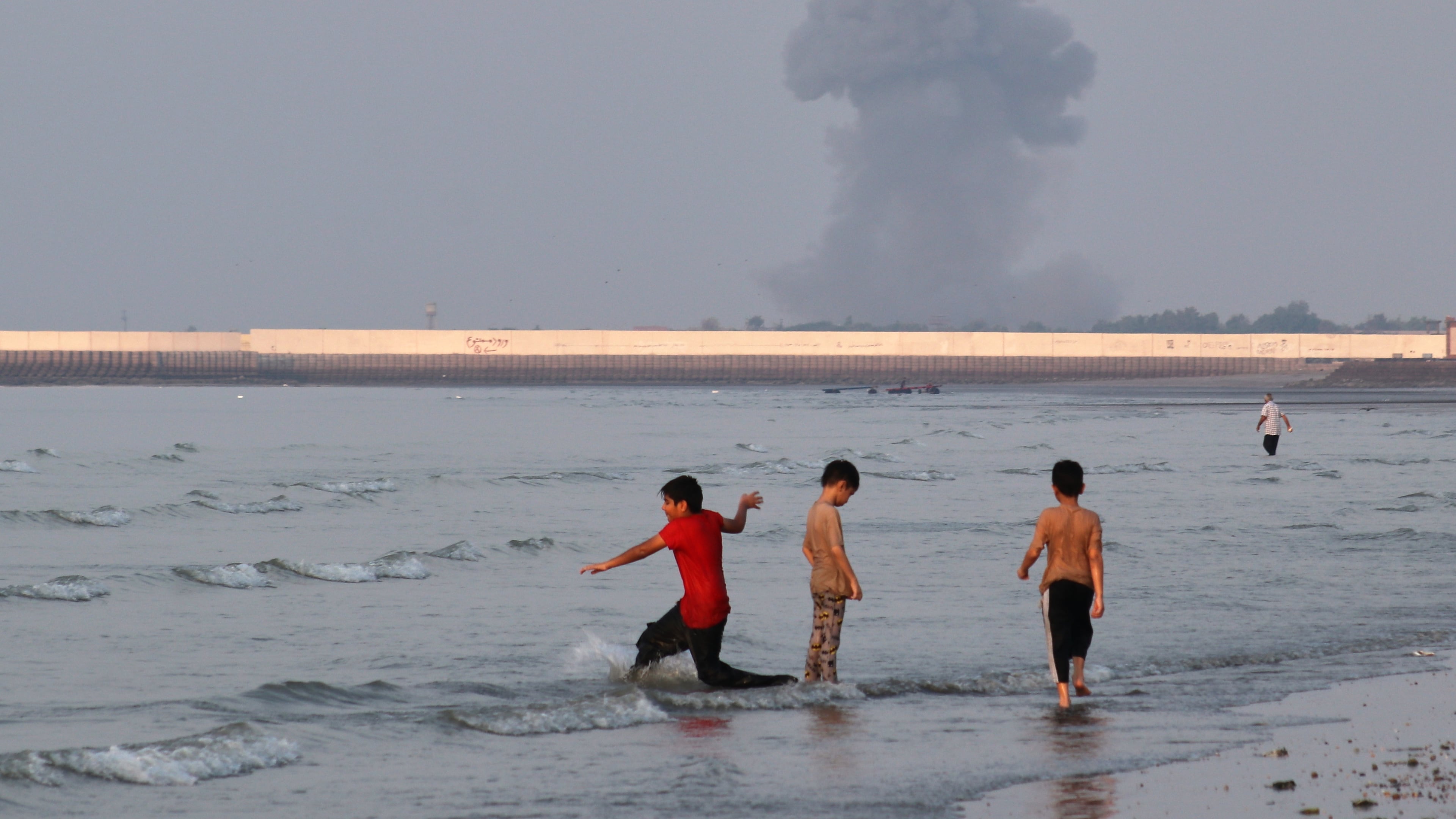 Three boys play in the shallow waters of the Strait of Hormuz, as a plume of smoke rises from an explosion in the background, off Bandar Abbas, Iran, Monday, July 13, 2026. (Razieh Poudat/ISNA via AP)