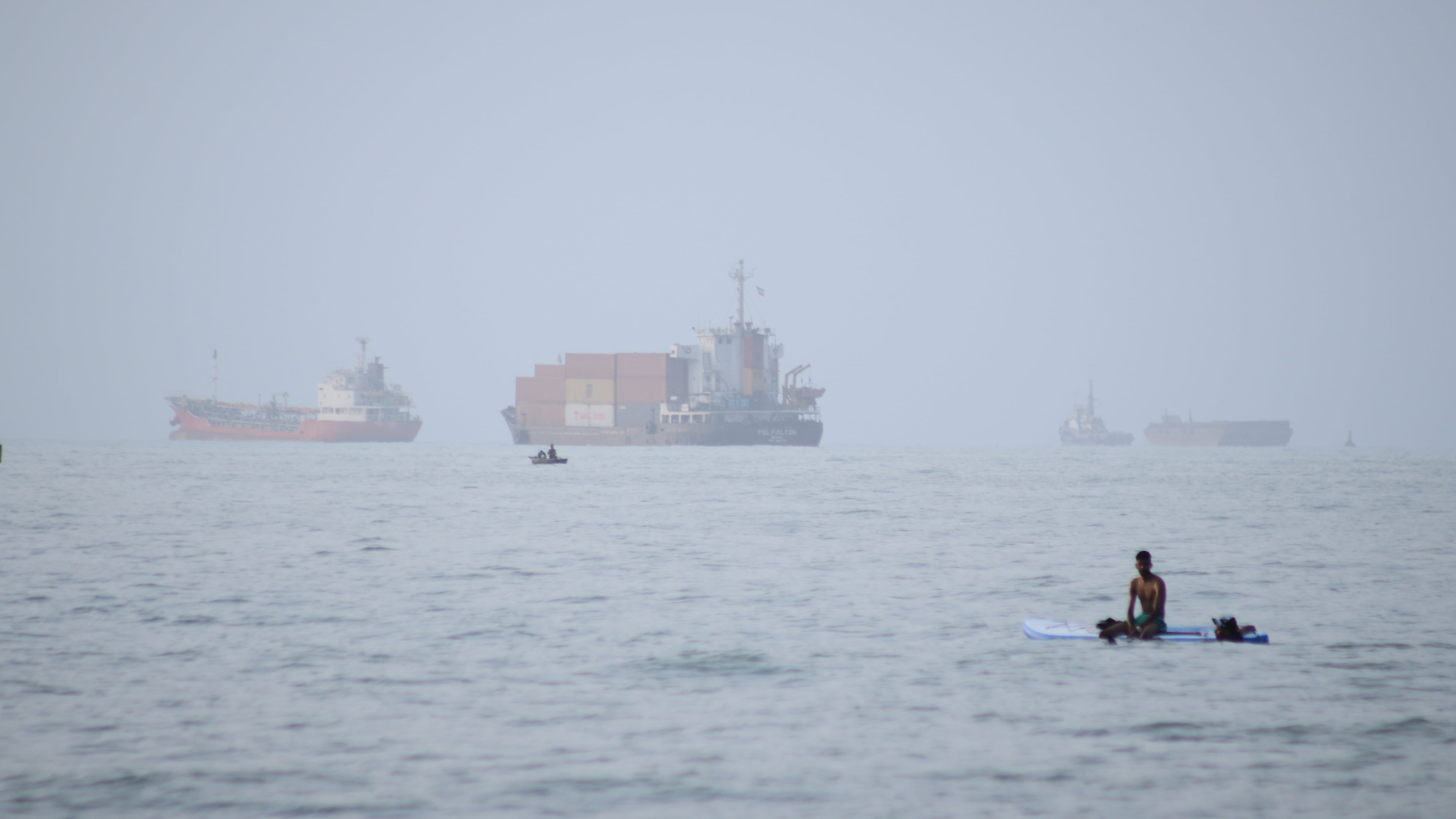 FILE - A man sits on a surfboard as a container ship and other commercial vessels appear anchored in the Strait of Hormuz off Bandar Abbas, Iran, Monday, July 27, 2026. (Razieh Poudat/ISNA via AP, File)