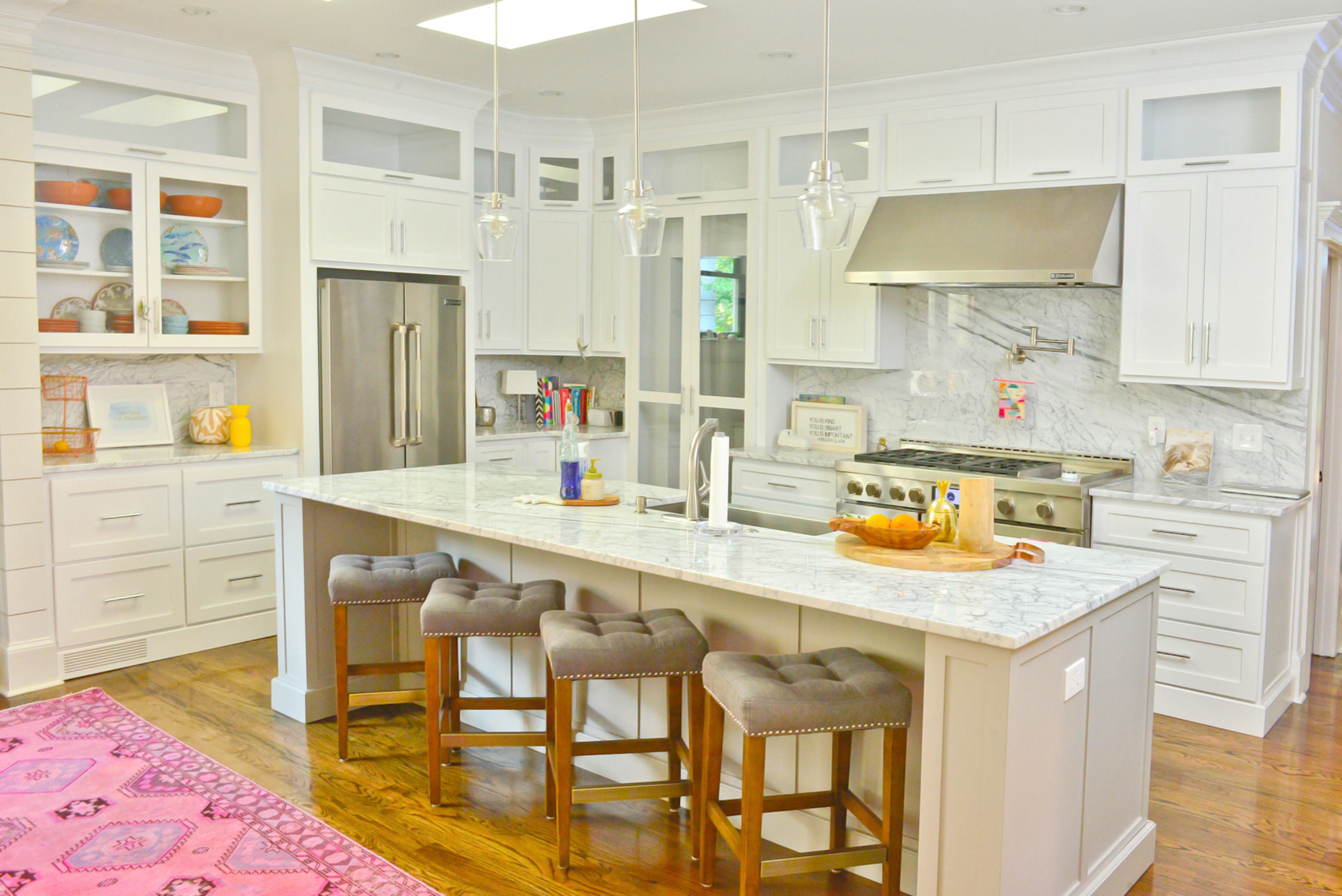Glass pendants hang above a wide Carrara marble-topped island in the center of the kitchen. Unexpected colors in the pink rug and pottery enliven the classic kitchen's bright white cupboards and marble counters. Plush barstools from Redefined Home Boutique in Atlanta provide comfortable seating.
