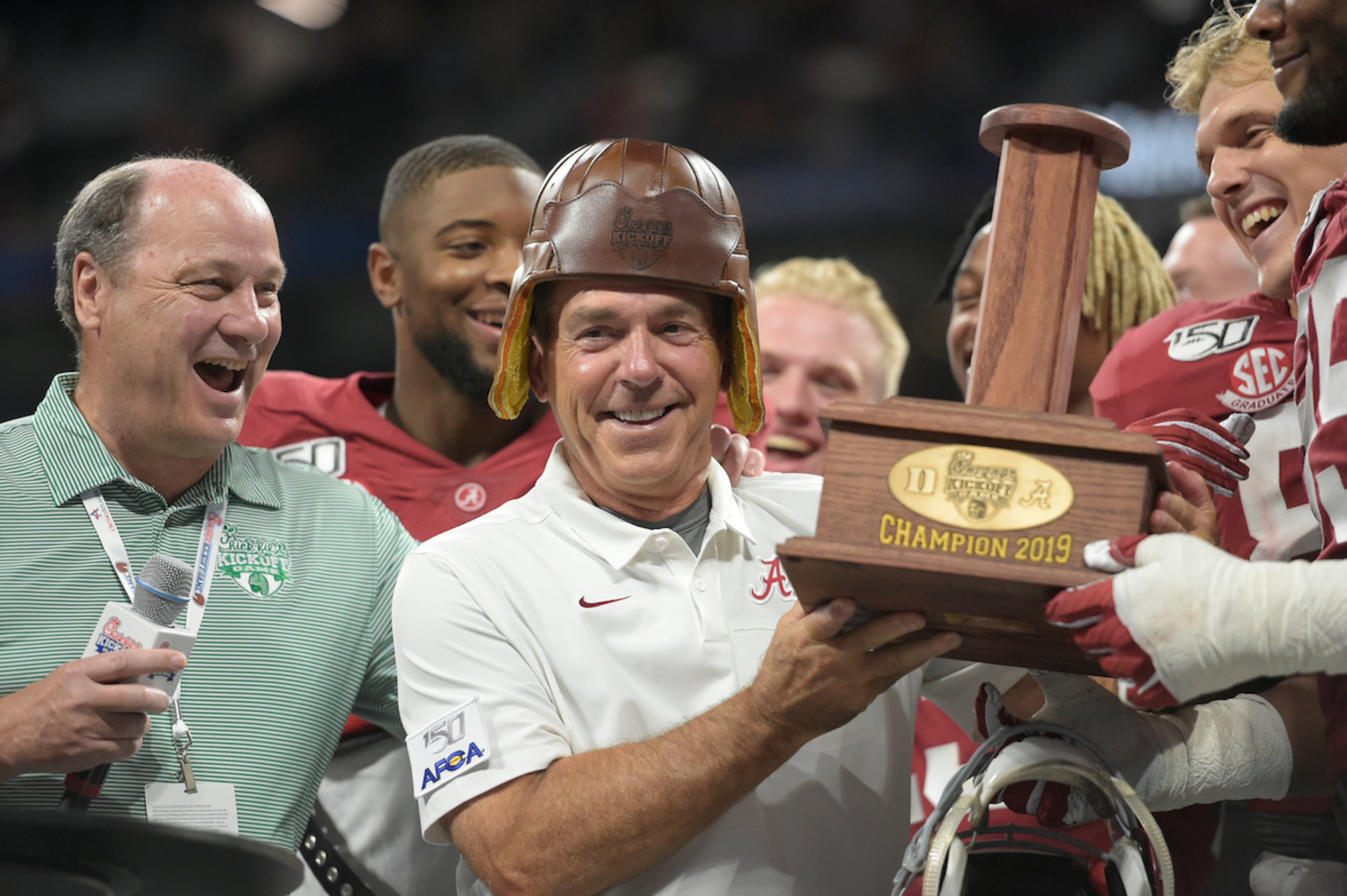 Peach Bowl Inc. CEO Gary Stokan (left) watches Alabama then-coach Nick Saban try on the "old leather helmet" from the Chick-fil-A Kickoff trophy at Mercedes-Benz Stadium in 2019. (Courtesy Vasha Hunt via Abell Images for Chick-fil-A Kickoff)