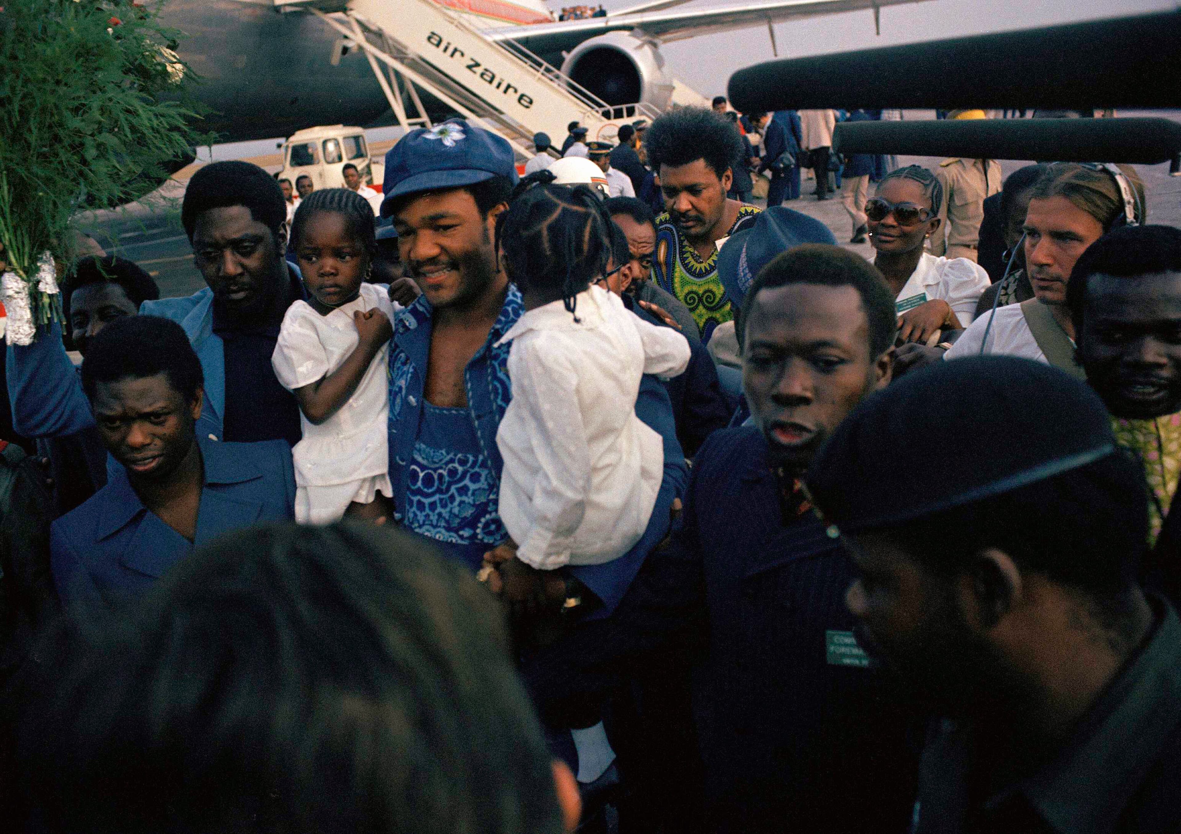FILE - This is a Sept. 12, 1974 file photo of heavyweight champion George Foreman as he carries two unidentified little girls as he arrives at Kinshasa, Zaire Airport, where the fight for the championship against Muhammad Ali will take place in October. Boxing promoter Don King is seen in background, centre, in green shirt. It was nearly 42 years ago that two men met just before dawn on Oct. 30, 1974, to earn $5 million in the Rumble in the Jungle. In one of boxing's most memorable moments, Muhammad Ali stopped the fearsome George Foreman to recapture the heavyweight title in the impoverished African nation of Zaire. (AP Photo, File)