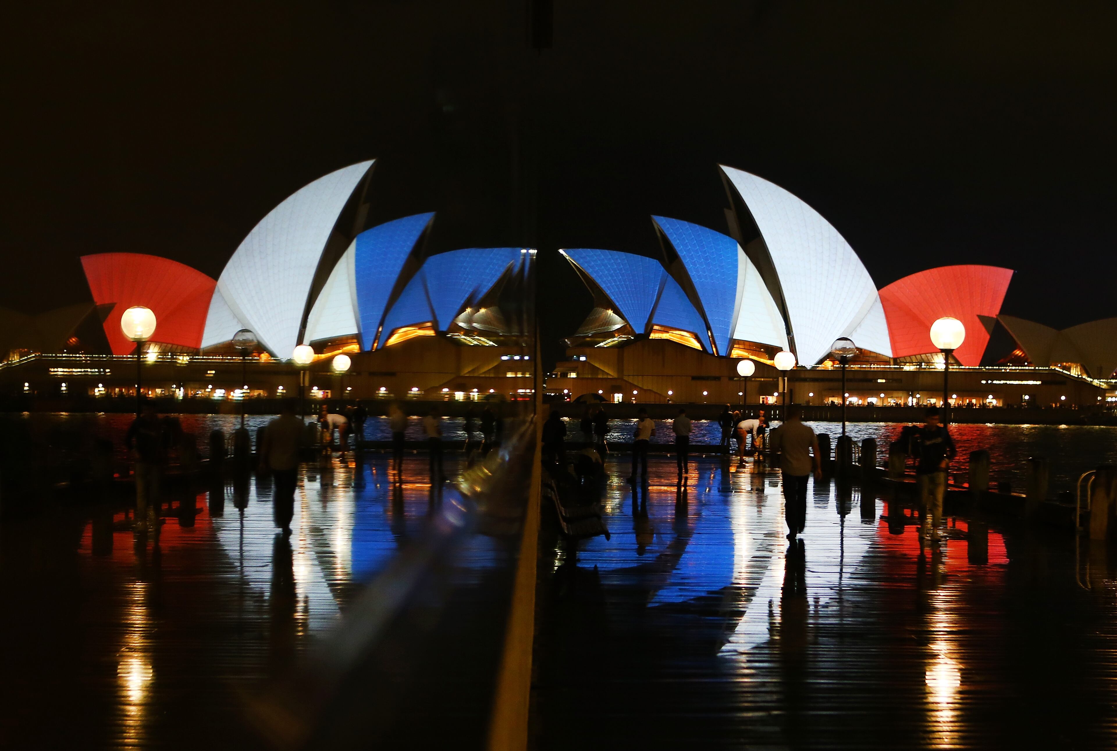 Pedestrians walk in front of a reflection of the Sydney Opera House as its sails are illuminated in the colours of the French flag on November 14, 2015 in Sydney, Australia. At least 120 people have been killed and over 200 are injured in Paris following a series of terrorist acts in the French capital on Friday. (Photo by Daniel Munoz/Getty Images)