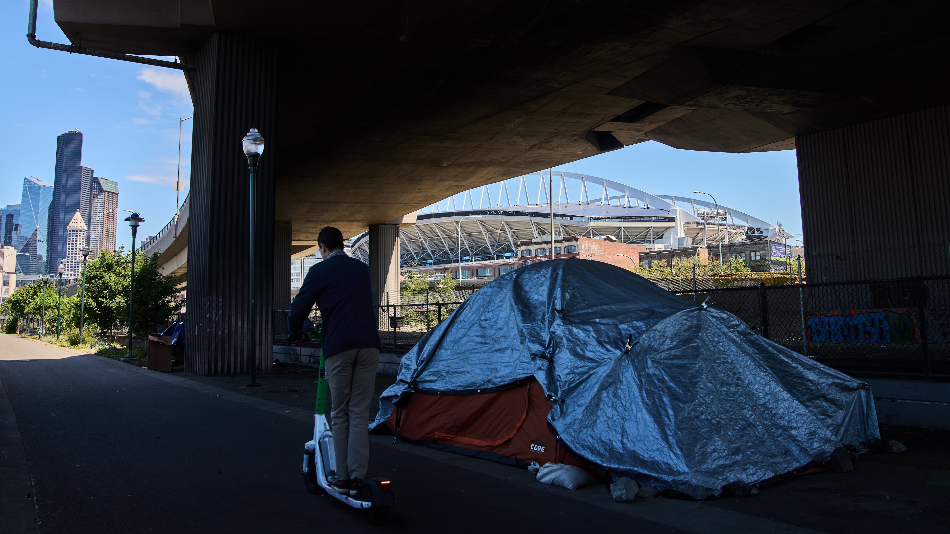 Seattle Stadium is seen in the background as a person rides a scooter past a series of tents on a trail near the stadium ahead of the 2026 FIFA World Cup soccer matches Wednesday, May 27, 2026, in downtown Seattle. (AP Photo/Lindsey Wasson)