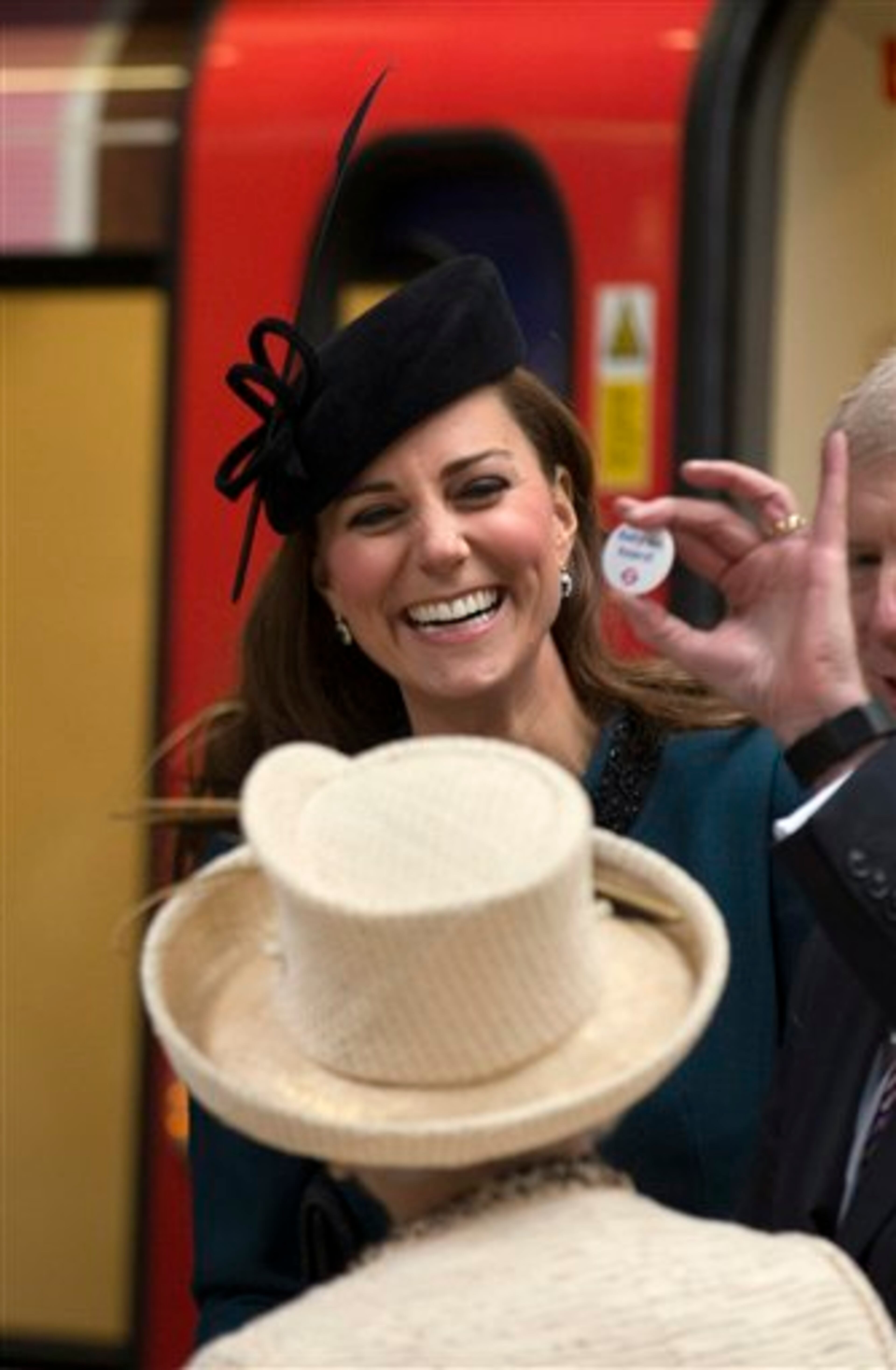 Britain's Kate, the Duchess of Cambridge, reacts as she is presented a badge that says "Baby on Board", normally available to pregnant passengers to encourage others to give them a seat, watched by Britain's Queen Elizabeth II, bottom, at Baker Street underground station in London, for a visit to mark the 150th anniversary of the London Underground, Wednesday, March 20, 2013. The Queen made her first public engagement in more than a week Wednesday after cancellations following her hospitalization for a stomach bug. The British head of state joined her husband Prince Philip and their granddaughter-in-law, Kate, for the event marking the 150th anniversary of London's sprawling subway system, affectionately known as the Tube. (AP Photo/Matt Dunham)