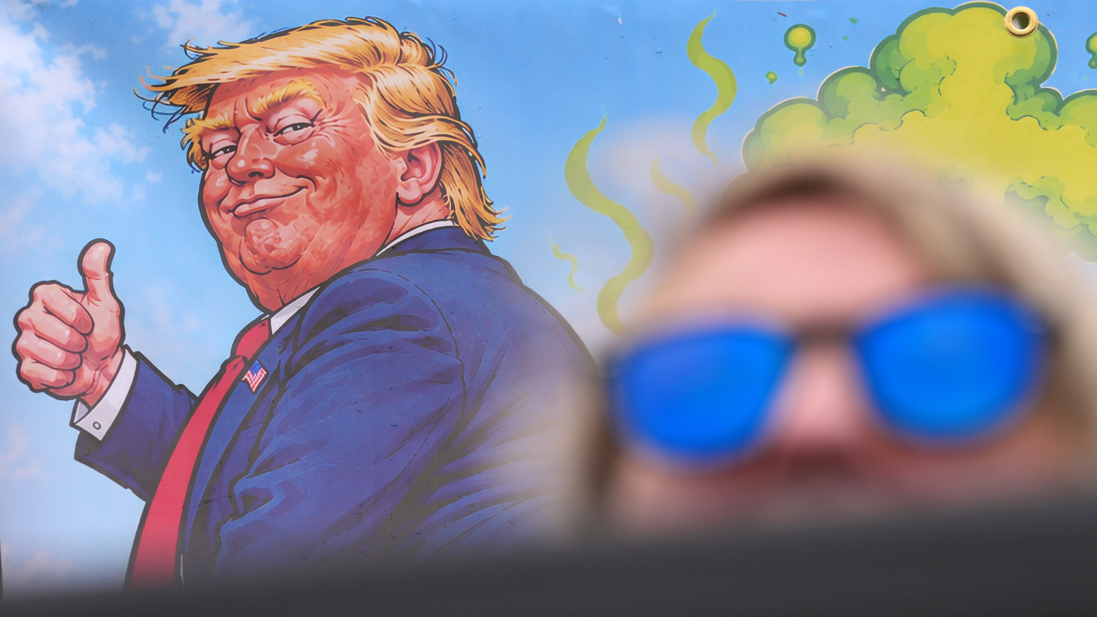 A protester wears sunglasses and sits in front of a poster depicting U.S. President Donald Trump during his visit to the Irish Open at the Trump International Golf Links course in Doonbeg, Ireland, Sunday, Sept. 13, 2026.(AP Photo/Kin Cheung)
