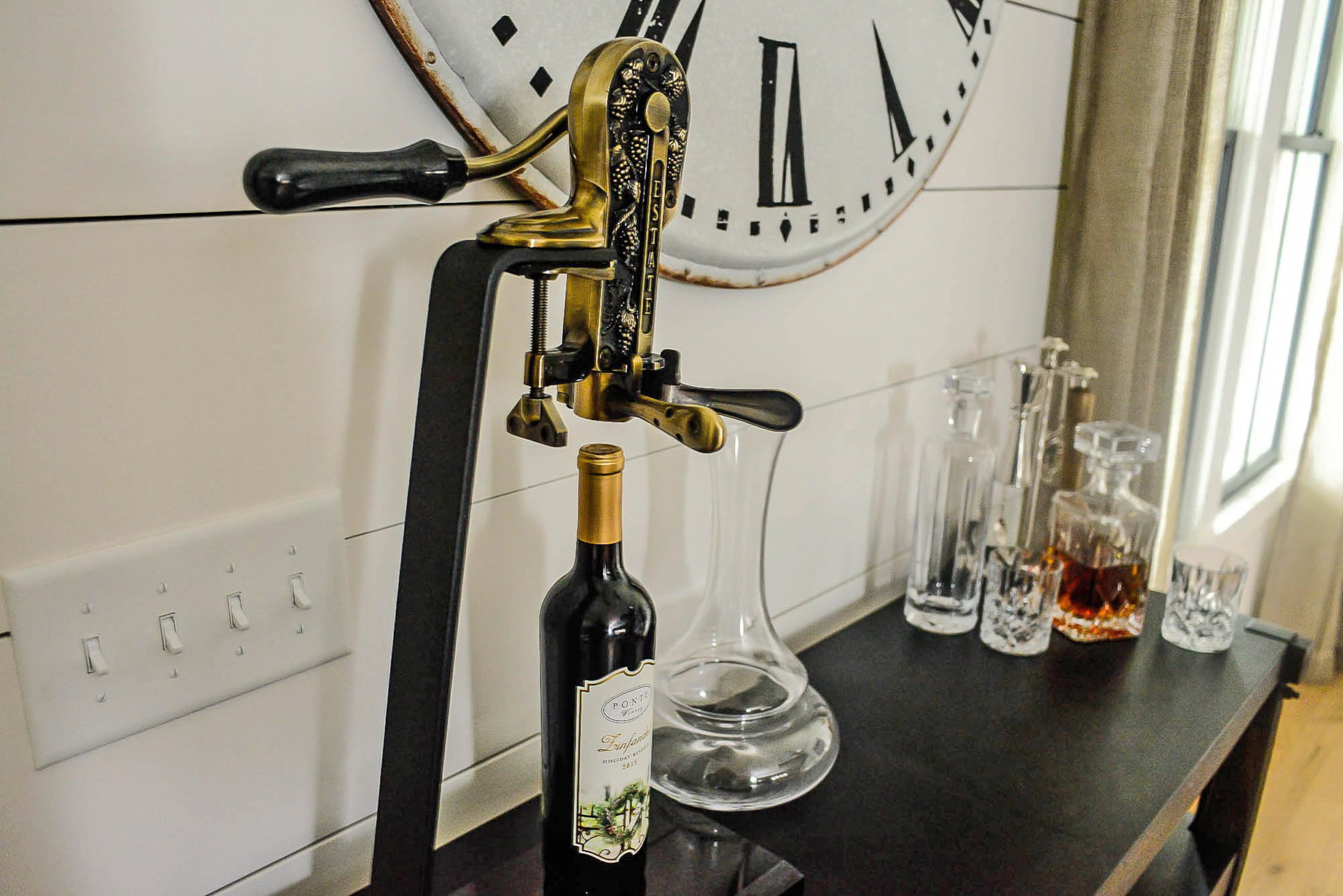 A vintage wine opener is attached to a rolling bar cart, which displays crystal brandy decanters. The oversized wall clock is from Urban Farmhouse.