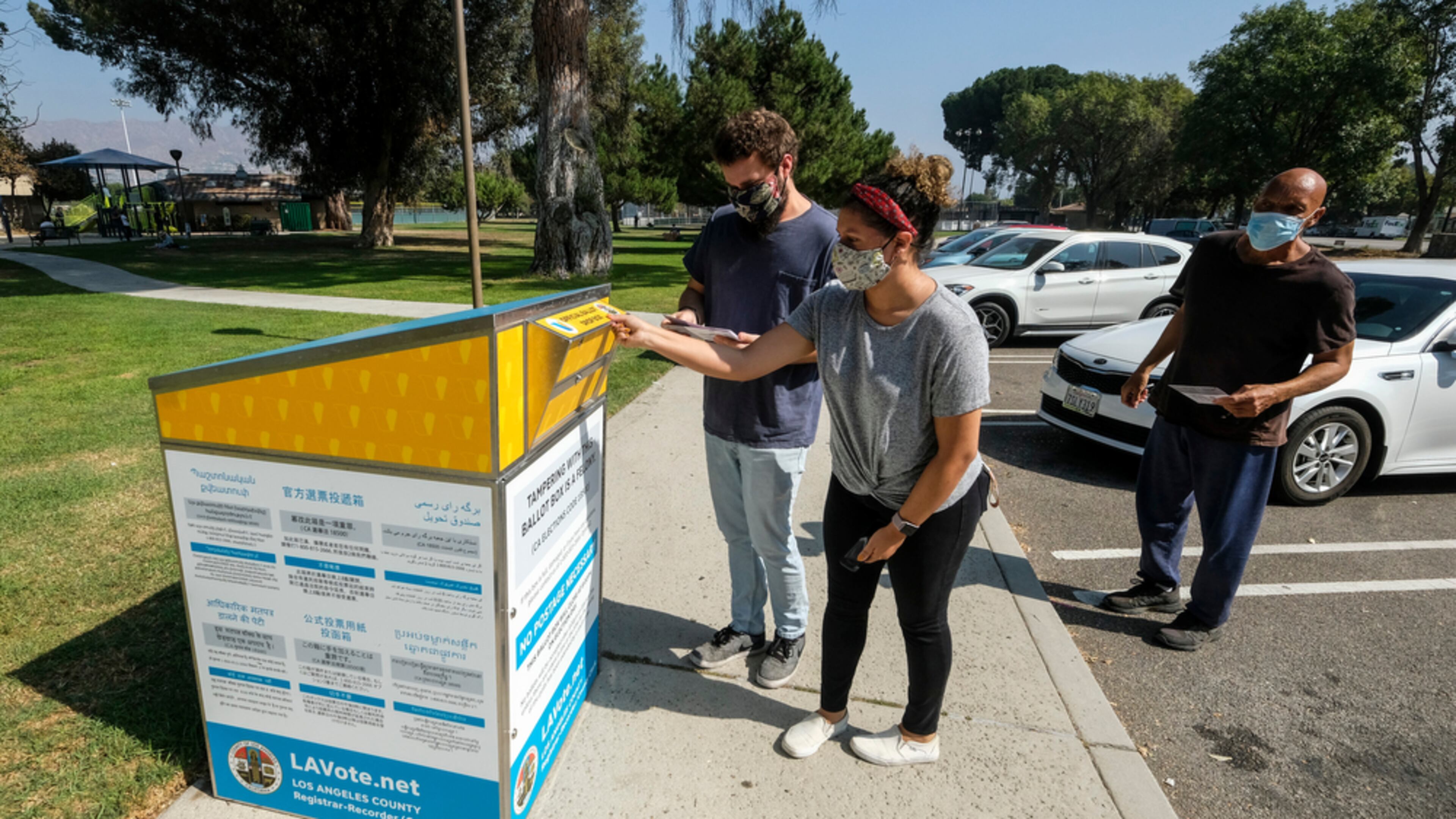 three people, a woman and two men, with face masks outside dropping voting ballots into a dropbox