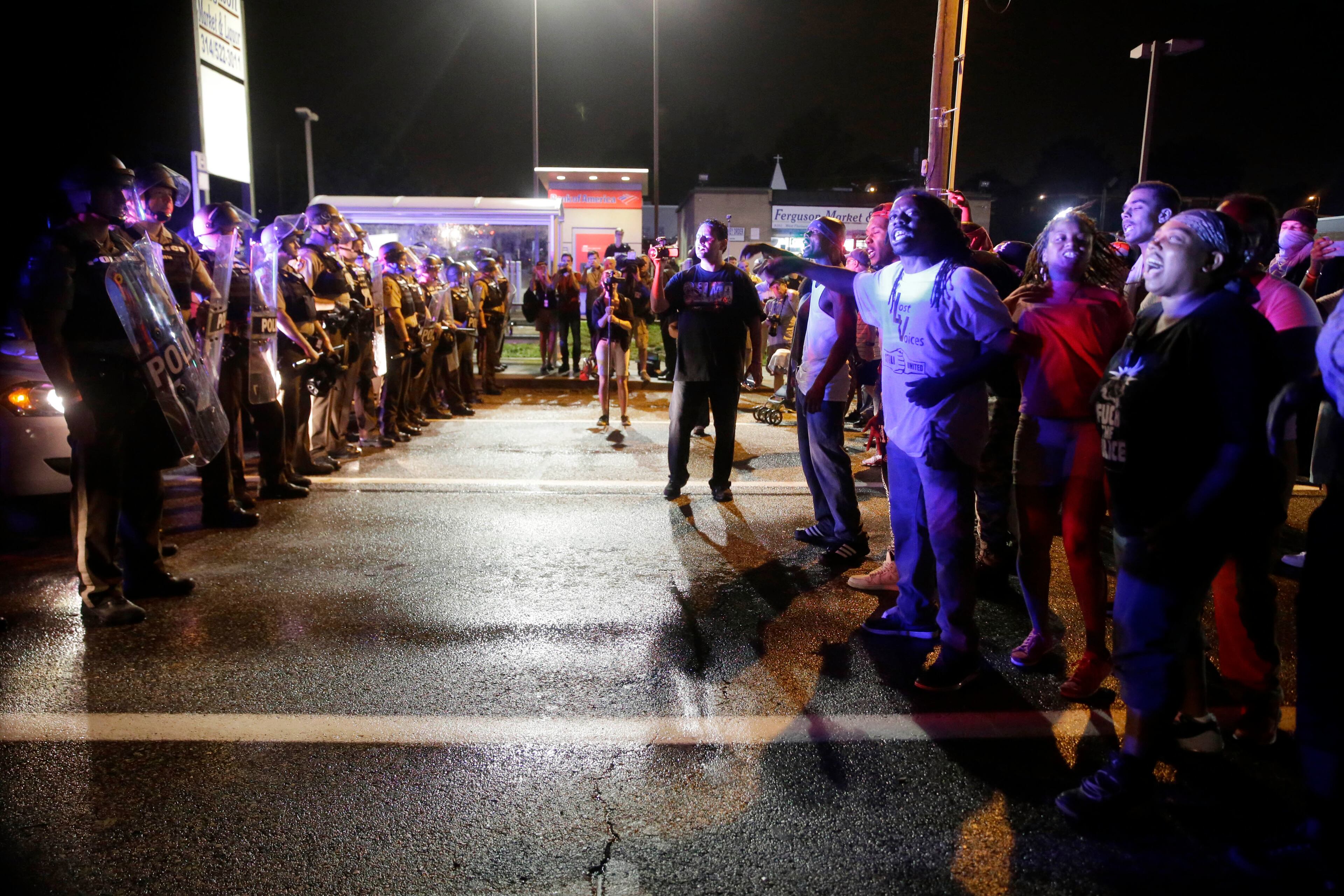 Protesters yell as police form a line across West Florissant Ave., Sunday, Aug. 9, 2015, in Ferguson, Mo., before shots were fired near the protest. The one-year anniversary of Michael Brown's death in Ferguson began with a march in his honor and ended with a protest that was interrupted by gunfire. (AP Photo/Jeff Roberson)