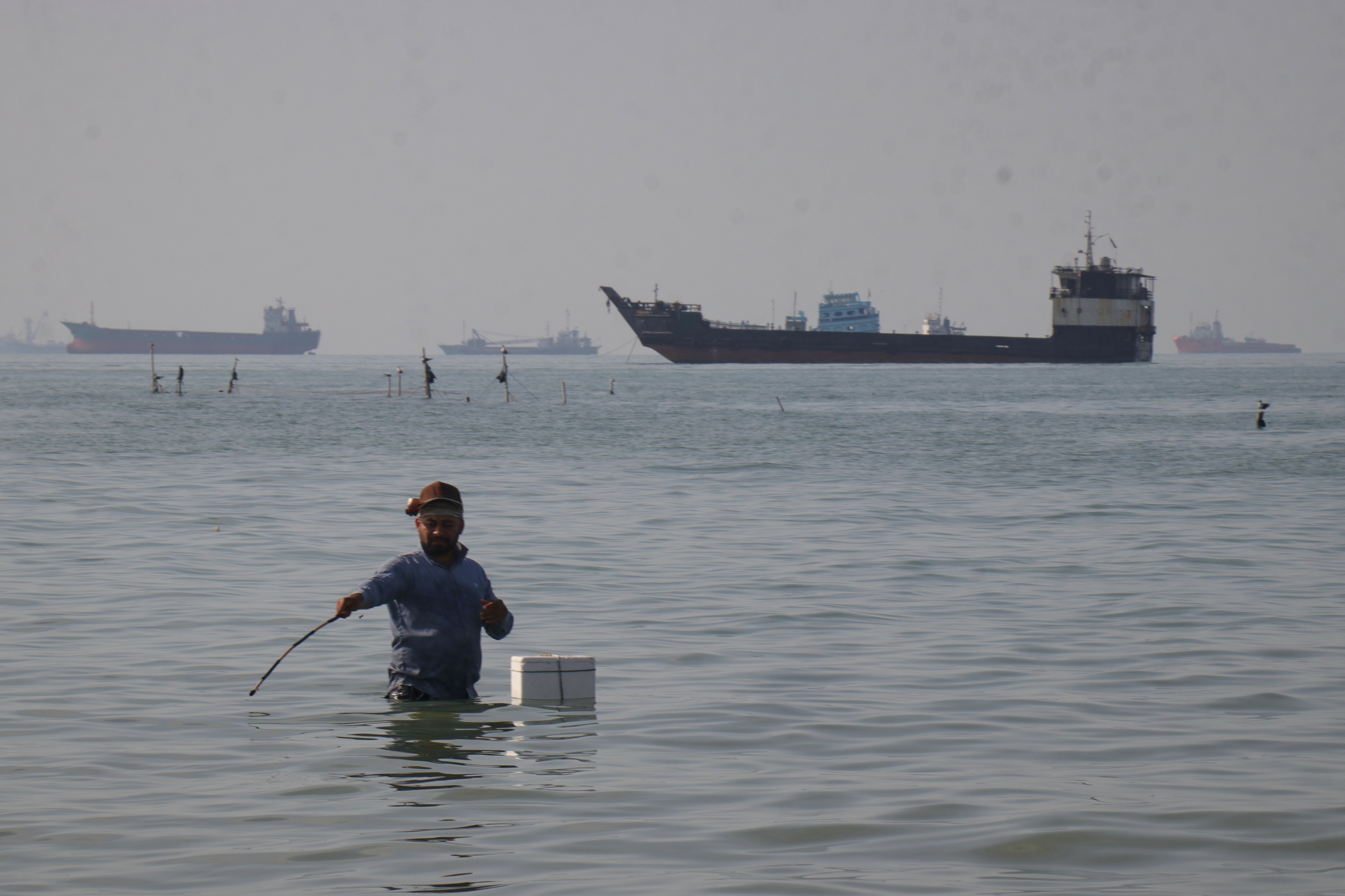 A man stands in the water, appearing to fish, as bulk carriers, cargo ships, and service vessels line the horizon in the Strait of Hormuz off Bandar Abbas, Iran, Monday, April 27, 2026.(Razieh Poudat/ISNA via AP)